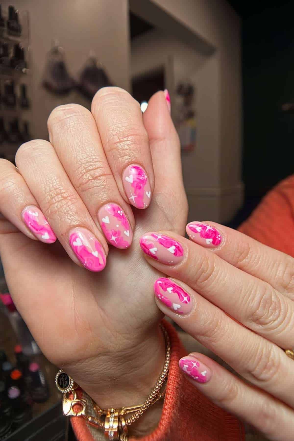 Hands with almond-shaped nails featuring pink and white abstract patterns and small heart designs. Person is wearing an orange long sleeve and gold bracelet.