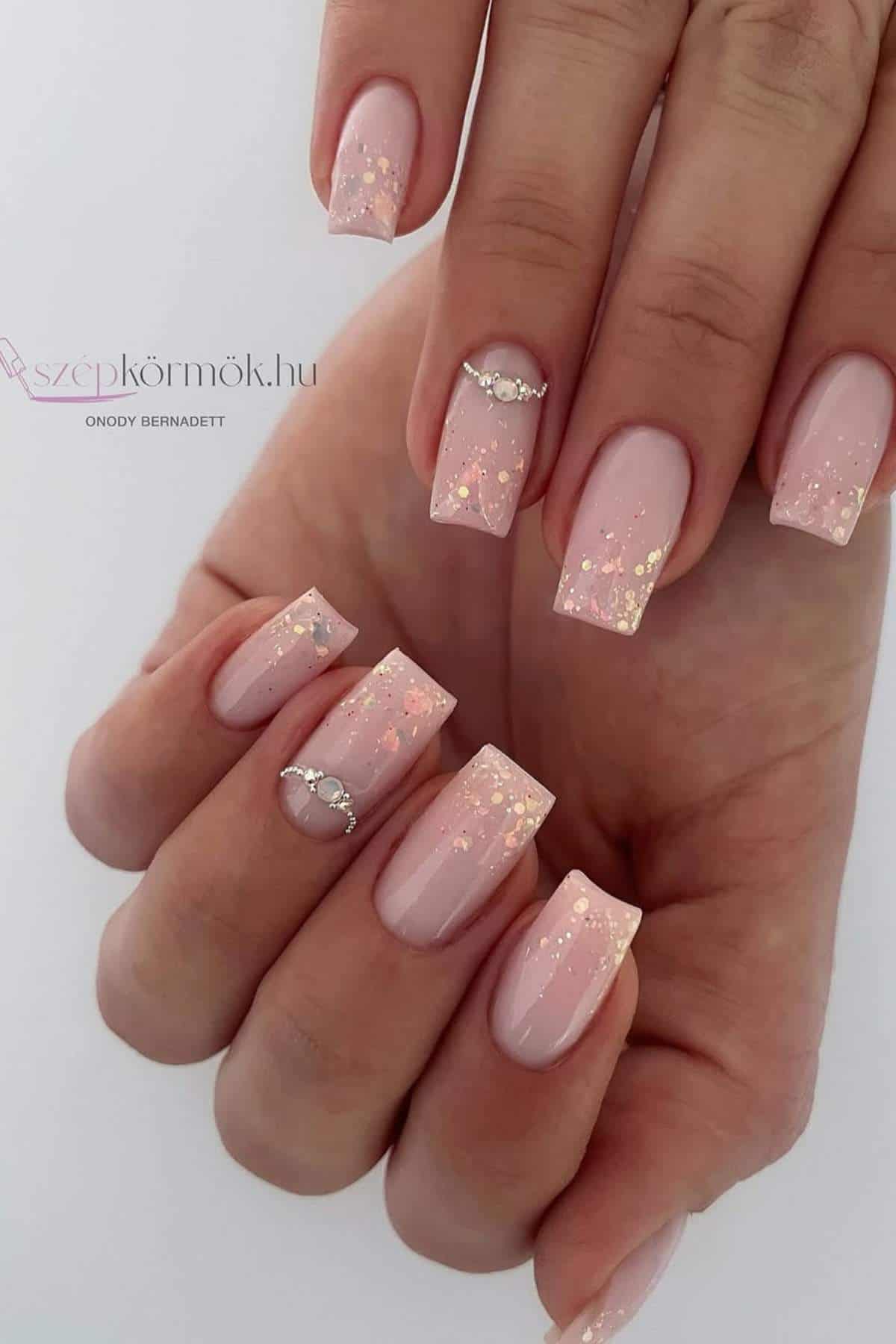 A hand with medium-length square nails painted light pink with gold glitter and rhinestone accents on two nails.