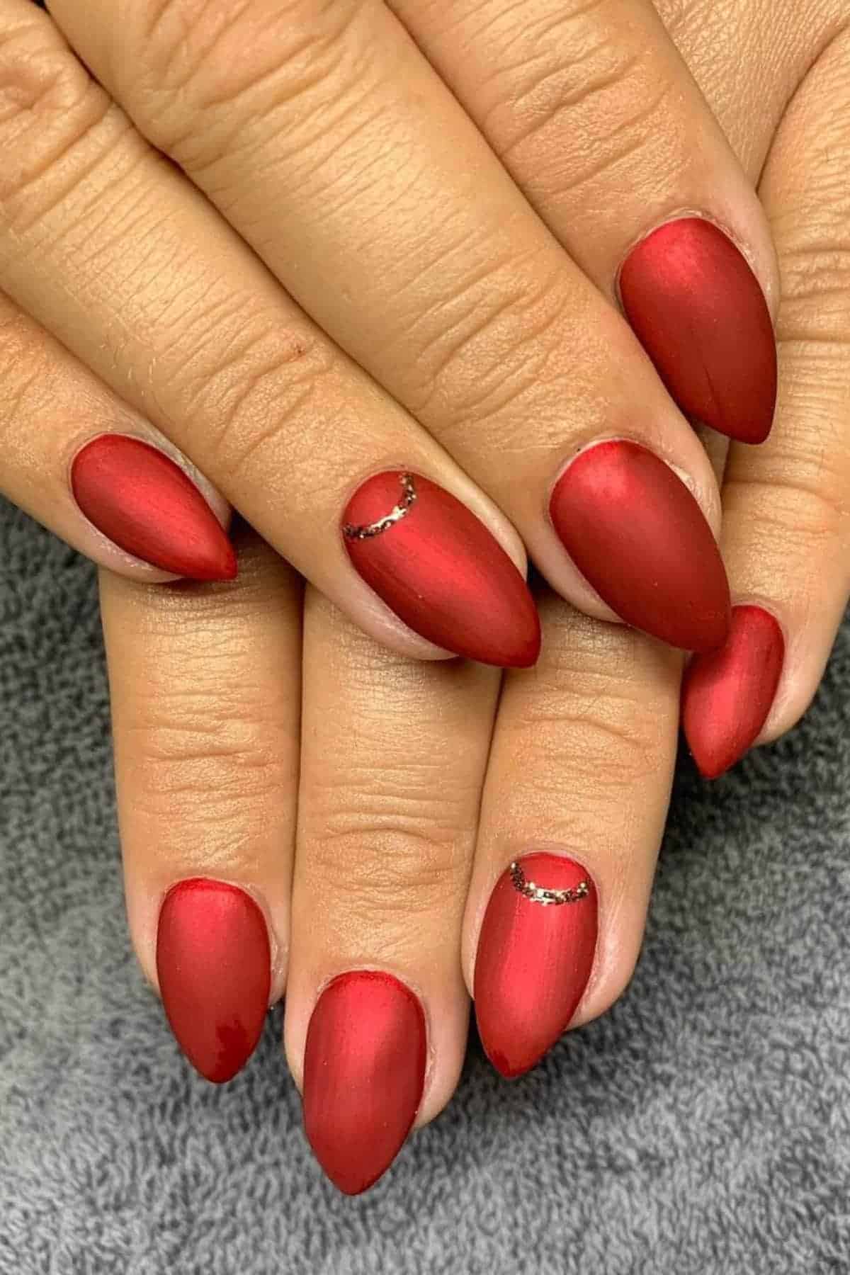 Hands with almond-shaped, matte red manicured nails, featuring a small rhinestone accent near the cuticle on two fingers, resting on a gray textured surface.