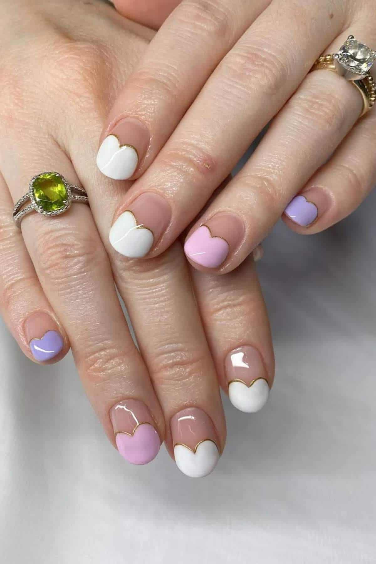 Hands with manicured nails featuring heart-shaped tips in white, pink, and lavender colors. The person wears two rings, one with a green stone and one with a clear stone.