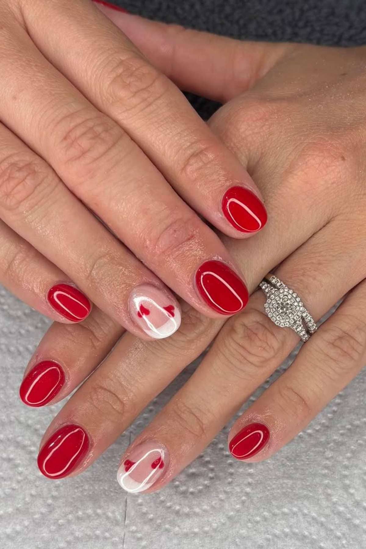 Hands with short, rounded red nails; ring finger on each hand has a clear base with small red heart designs. Person is wearing a diamond ring.