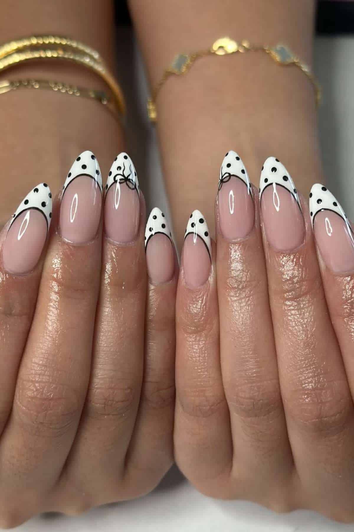 Hands with almond-shaped nails featuring white French tips, black polka dots, and thin black accent lines. The person is wearing gold bracelets.