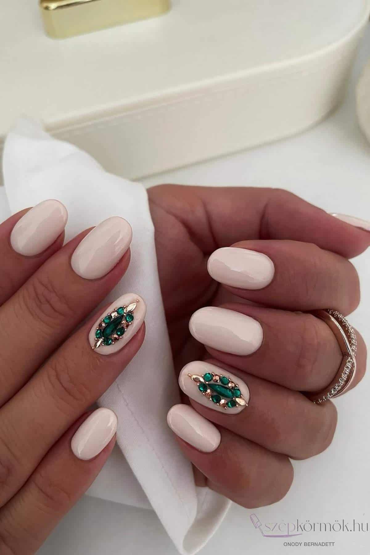 A hand with short, almond-shaped nails painted in pale pink polish, featuring green gemstone and gold bead accents on the ring fingernails.