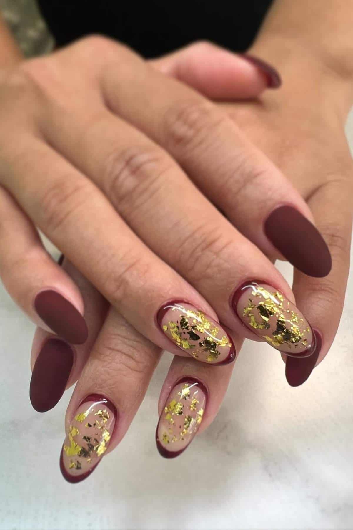 Hands with long, almond-shaped nails; most are painted matte deep red, while two nails on each hand have gold foil accents on a sheer base.