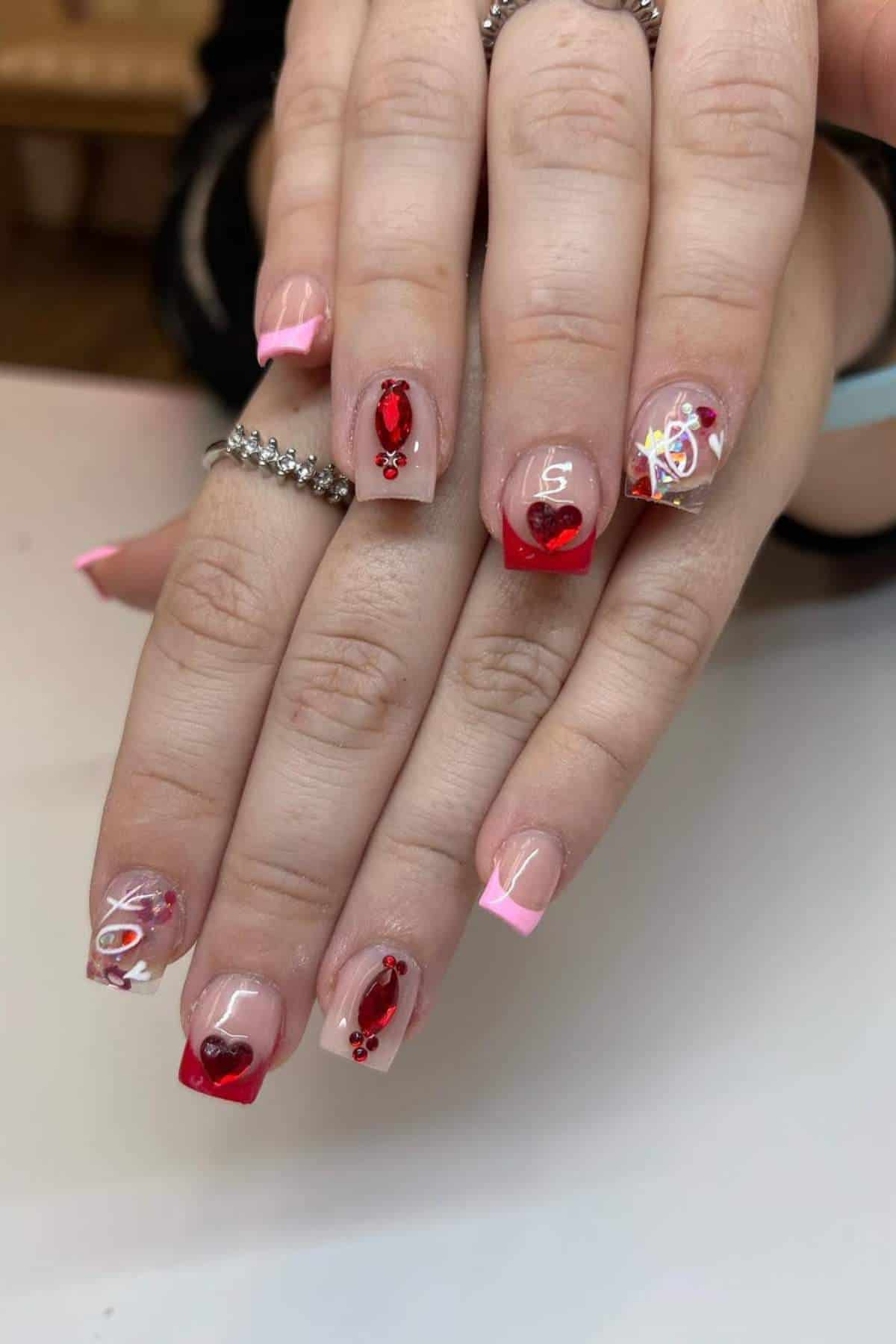 Hands with manicured nails featuring pink French tips and red heart-themed nail art, rhinestones, and decorative designs on a white surface.