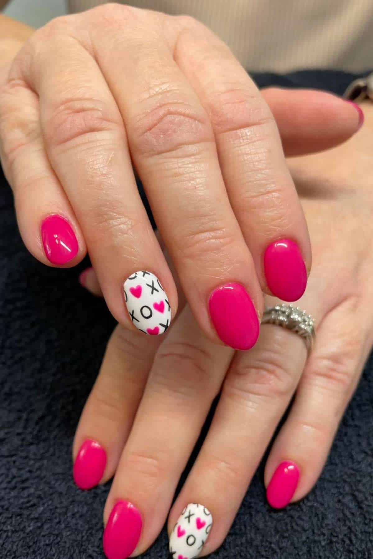 Hands with bright pink manicured nails; ring fingers feature a white base with black Xs and Os and pink hearts as accent nail art.