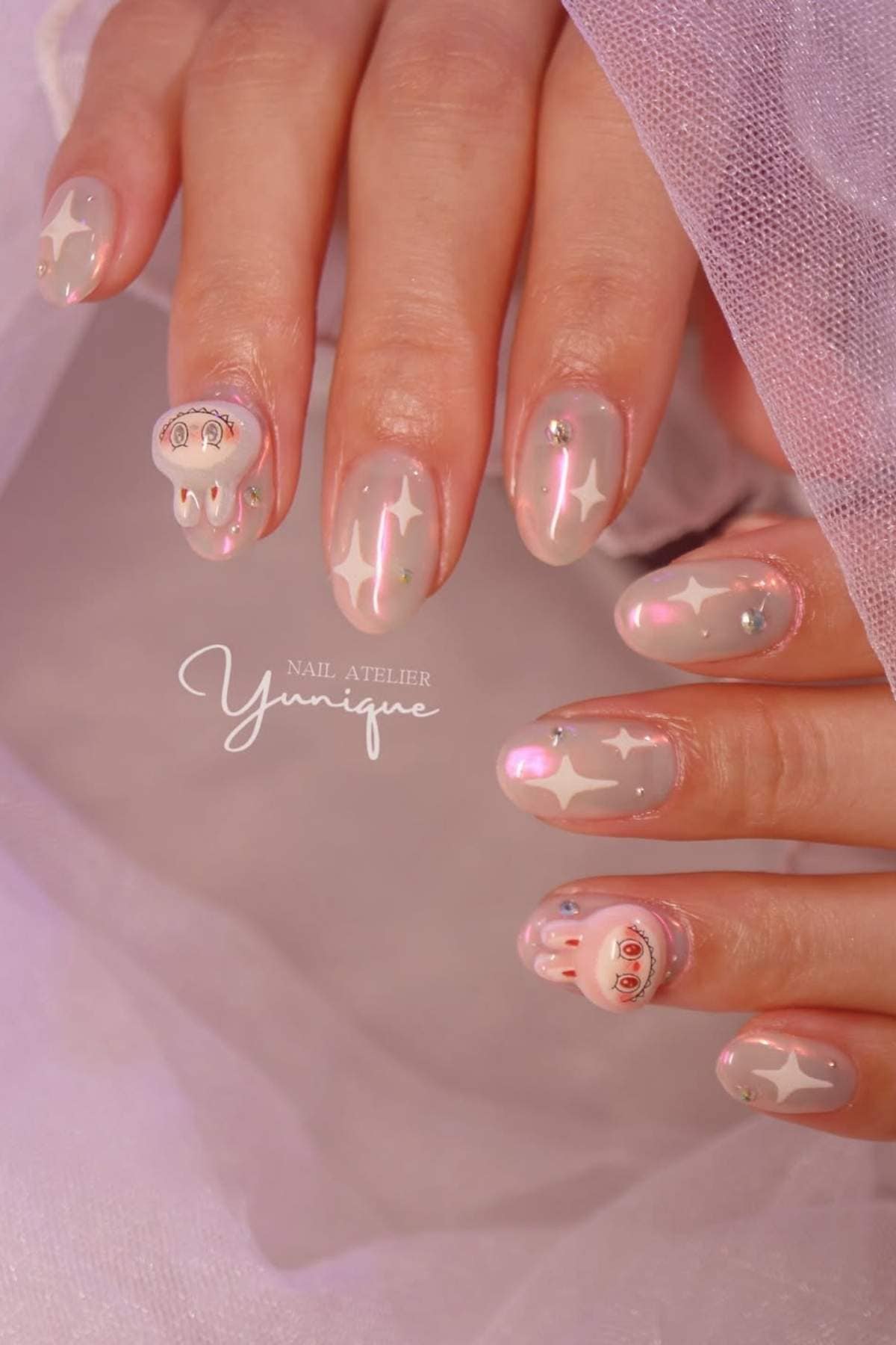 Hands with light pink manicured nails featuring white star designs, small rhinestones, and cartoon-like faces on two nails, shown against a soft fabric background.