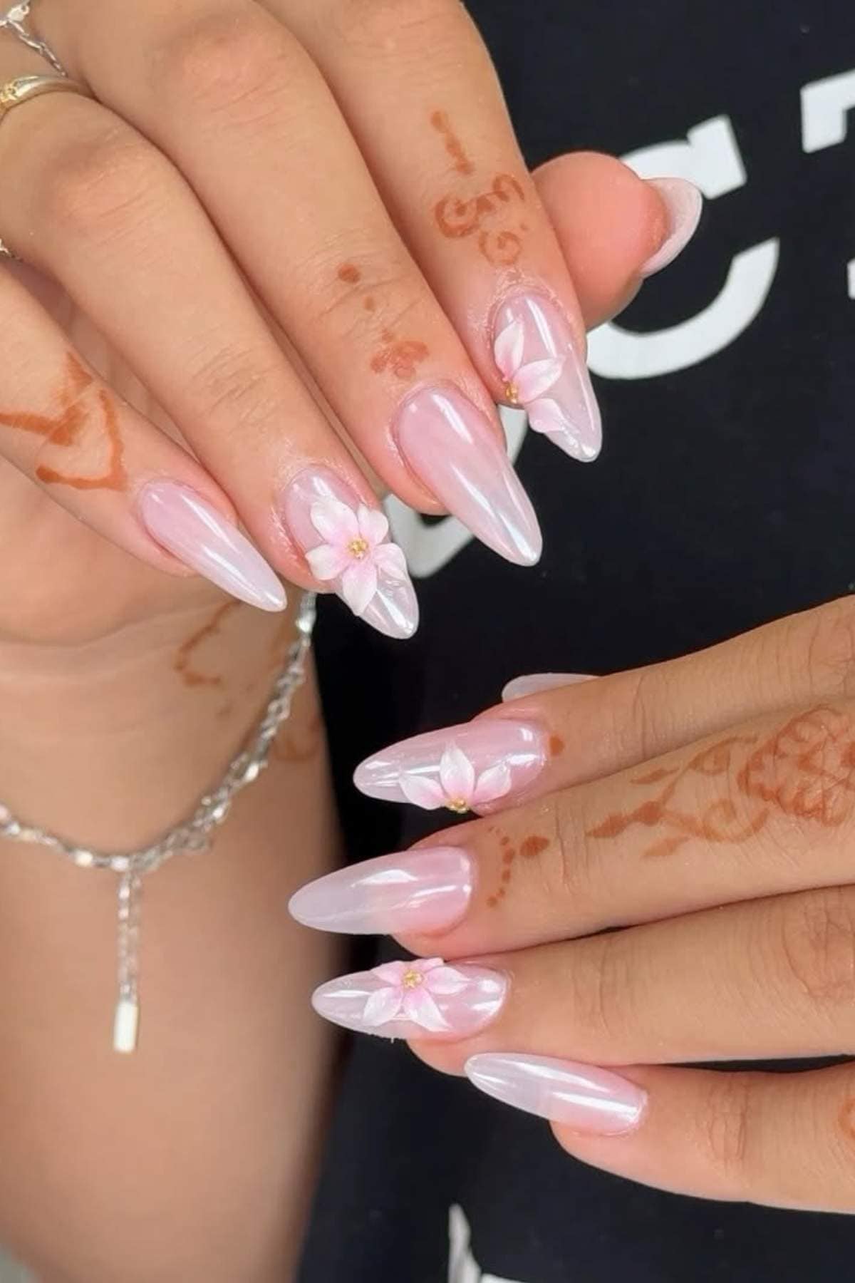 Hands with long, almond-shaped, glossy pale pink nails featuring 3D white floral designs and small gold accents; henna patterns are visible on the fingers.