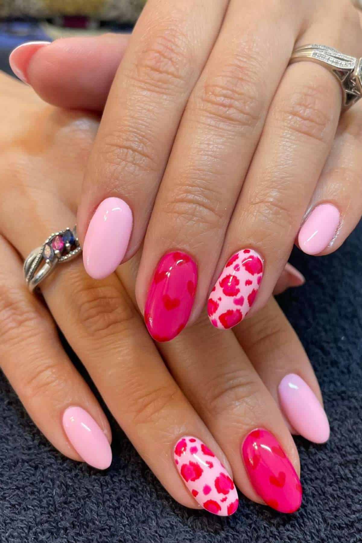 Hands with light pink manicured nails; two accent nails feature red and pink leopard spots and heart designs. Silver rings are worn on two fingers.