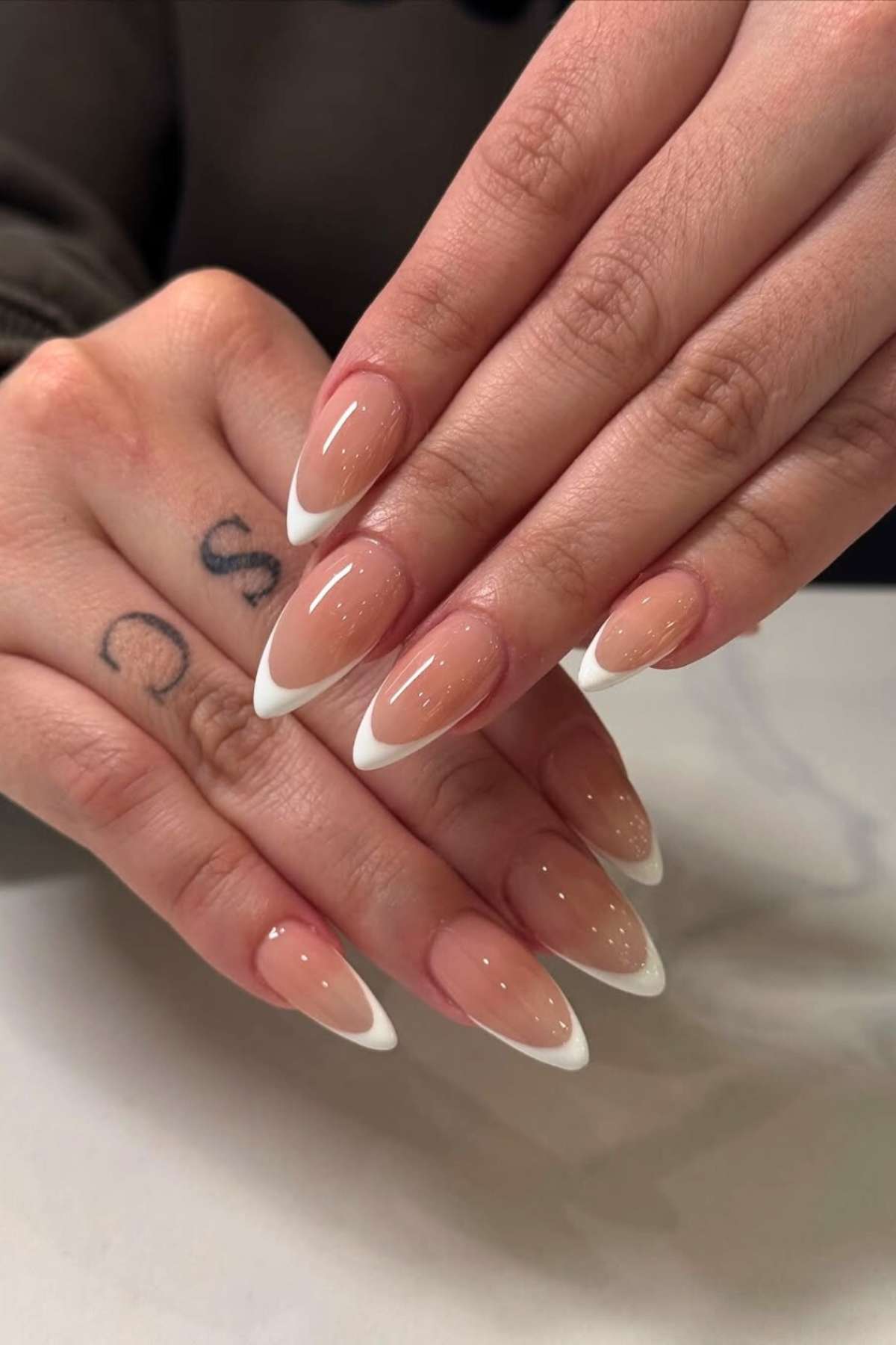 A close-up of hands with almond-shaped nails featuring a nude base and white French tips. One finger has a small “S” tattoo on the side.