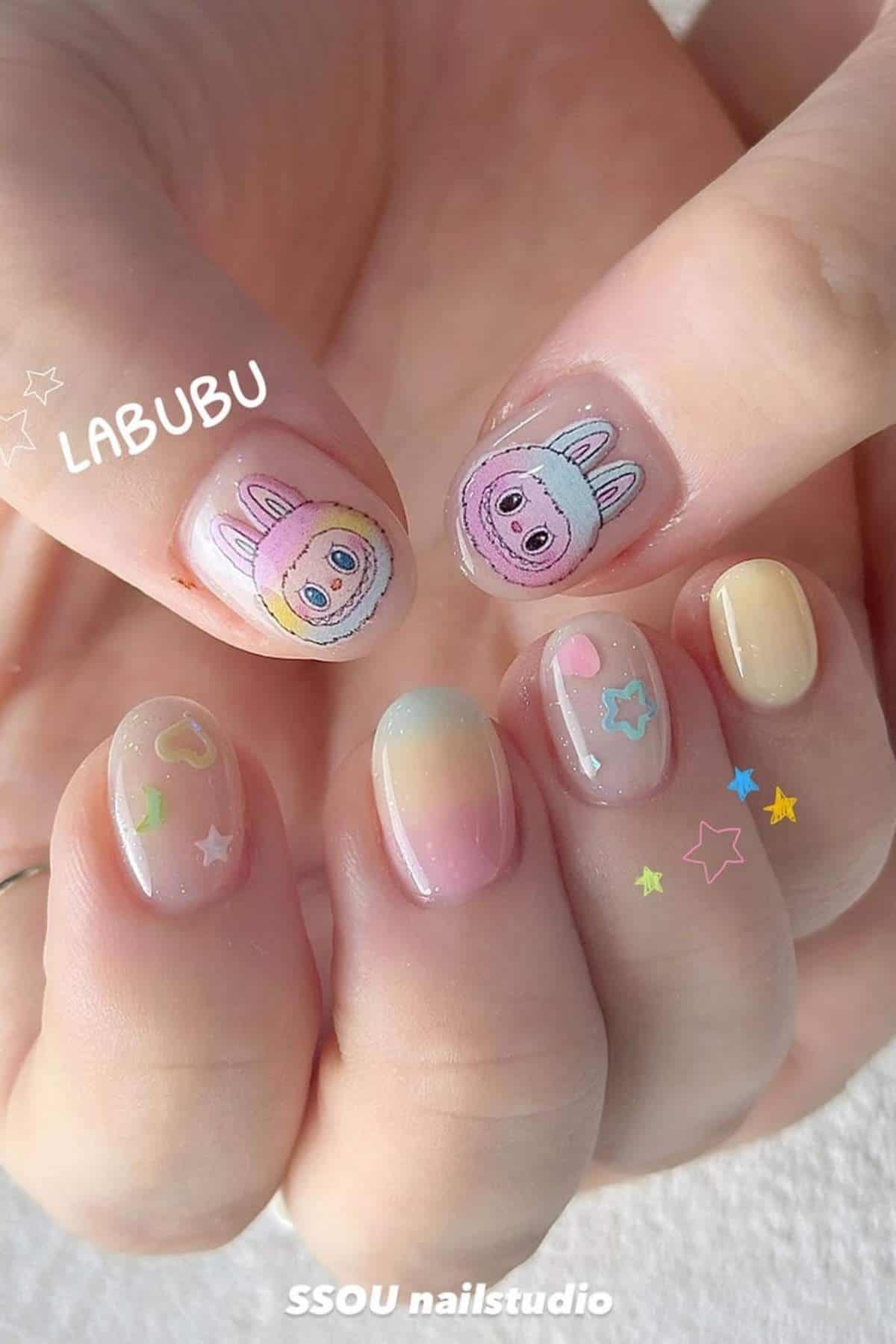 A hand with pastel gradient nails, two featuring cartoon animal faces labeled "LABUBU," and others decorated with small heart and star designs.