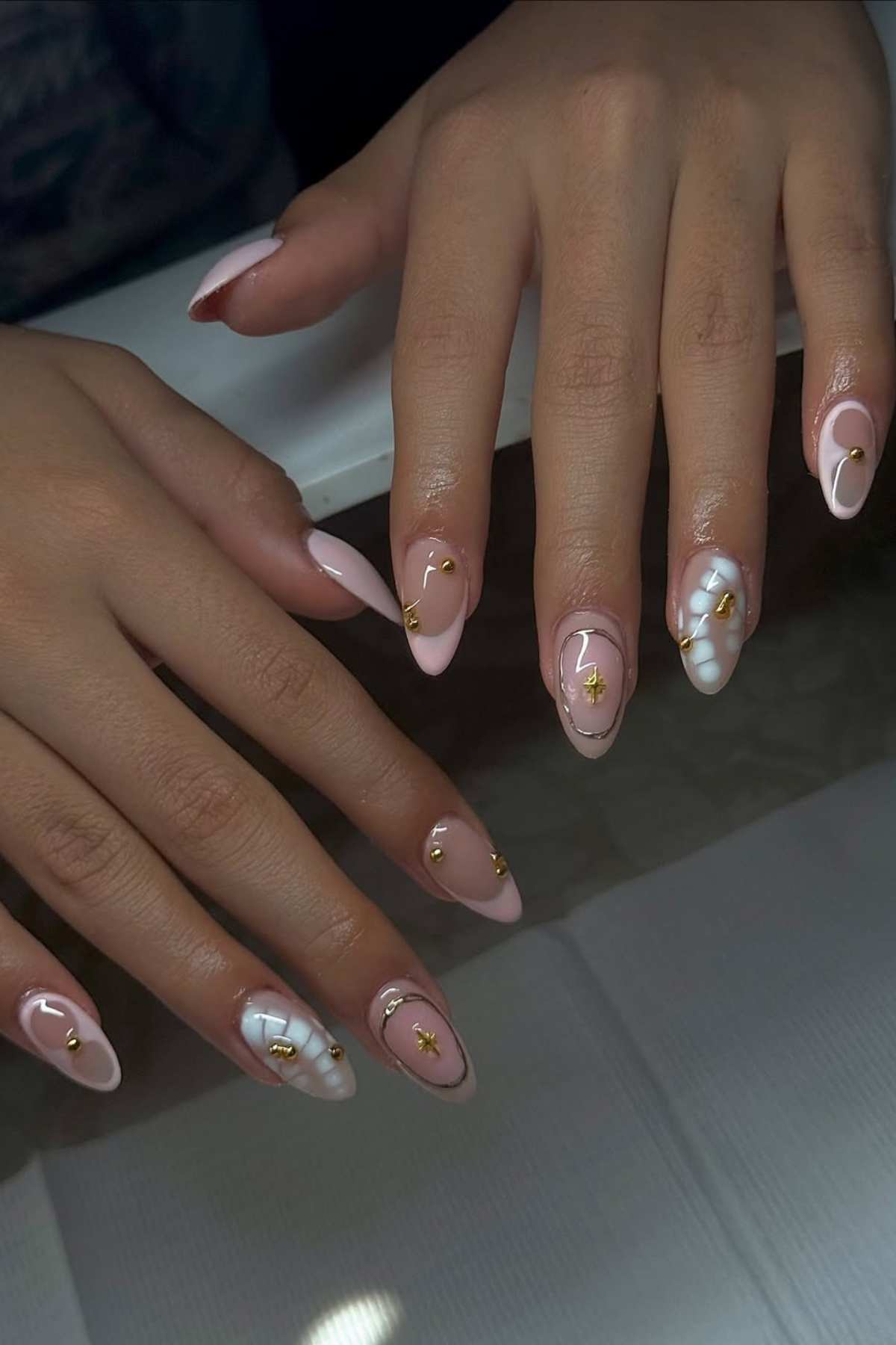 Two hands with almond-shaped nails painted in pale pink and white, decorated with gold accents and small gold charms.