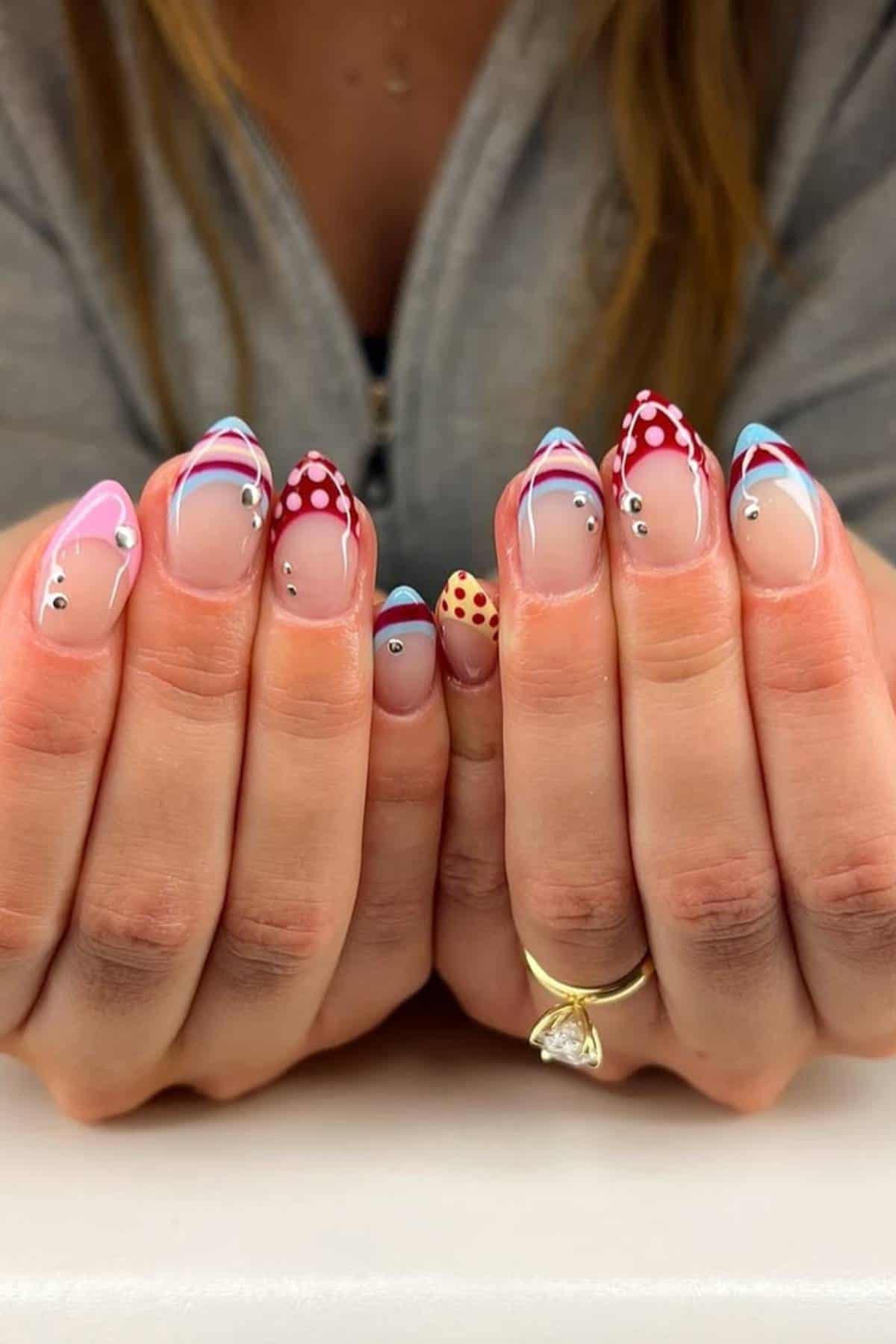 Close-up of hands with manicured nails featuring multicolored tips, red polka dots, and small rhinestone accents on a nude base.
