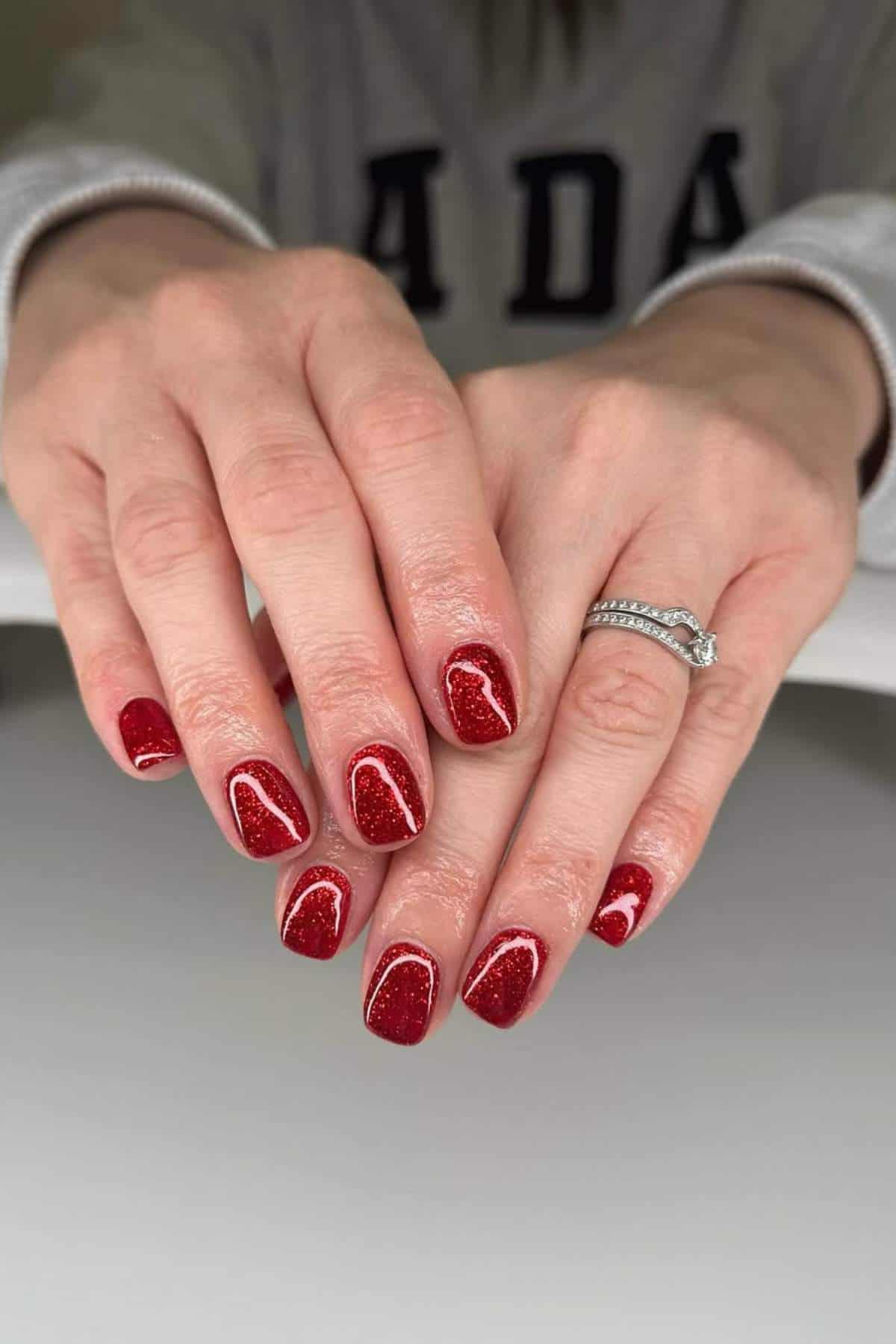 A person's hands with shiny red glitter nail polish and white outlines on short nails, wearing a silver ring with a clear stone, resting on a white surface.