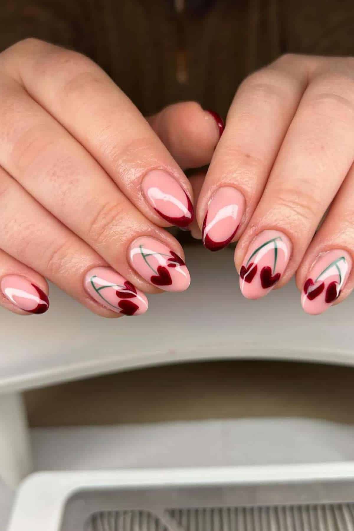 Hands with almond-shaped nails featuring a pink base and red cherry designs with green stems on each nail.