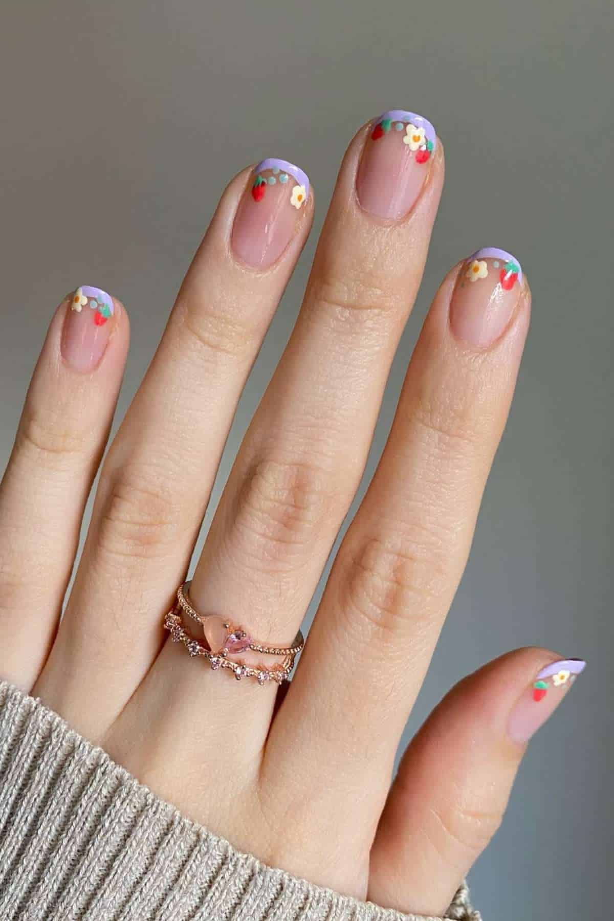 A hand with light skin and two rings displays nails with a clear base and colorful floral French tips.