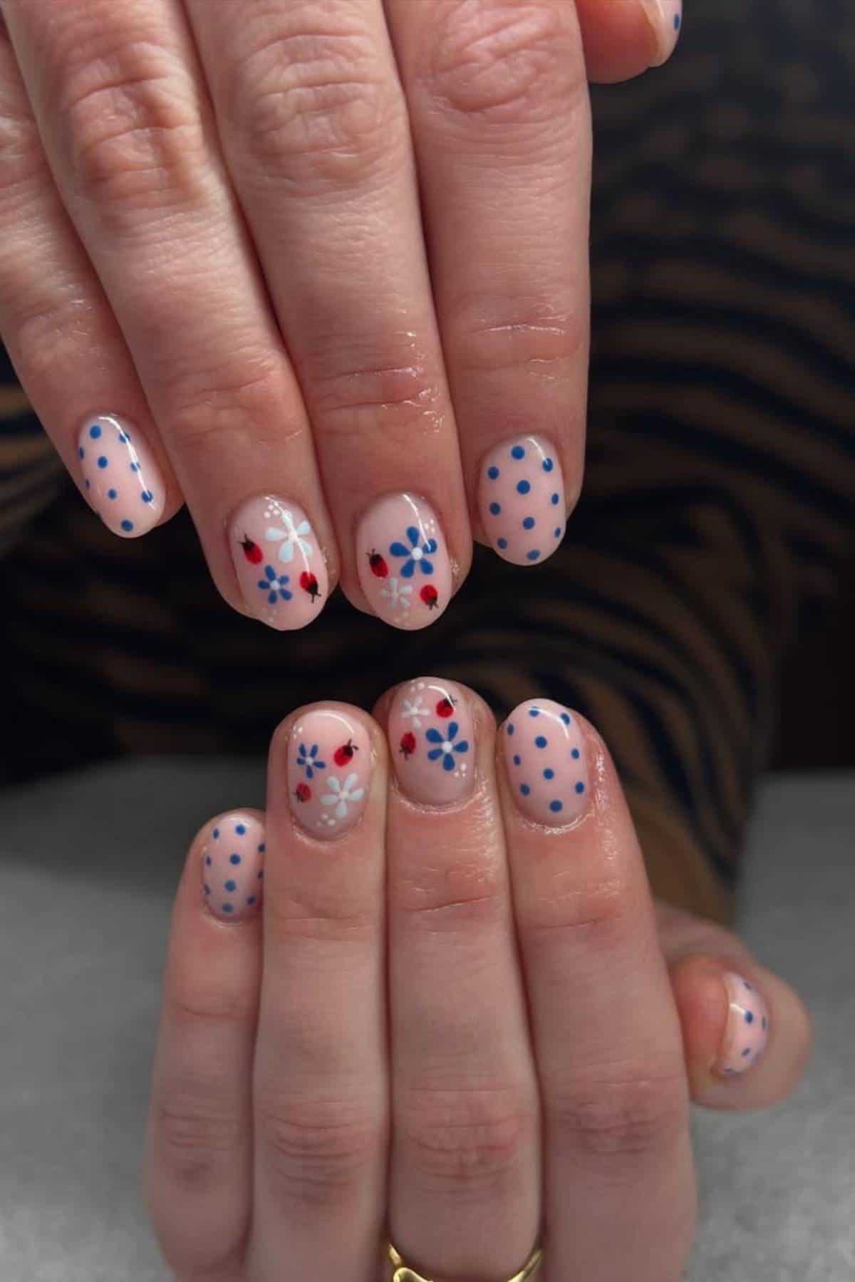 Close-up of hands with nails painted in a nude color, featuring blue polka dots and red, blue, and white flower designs on each nail.