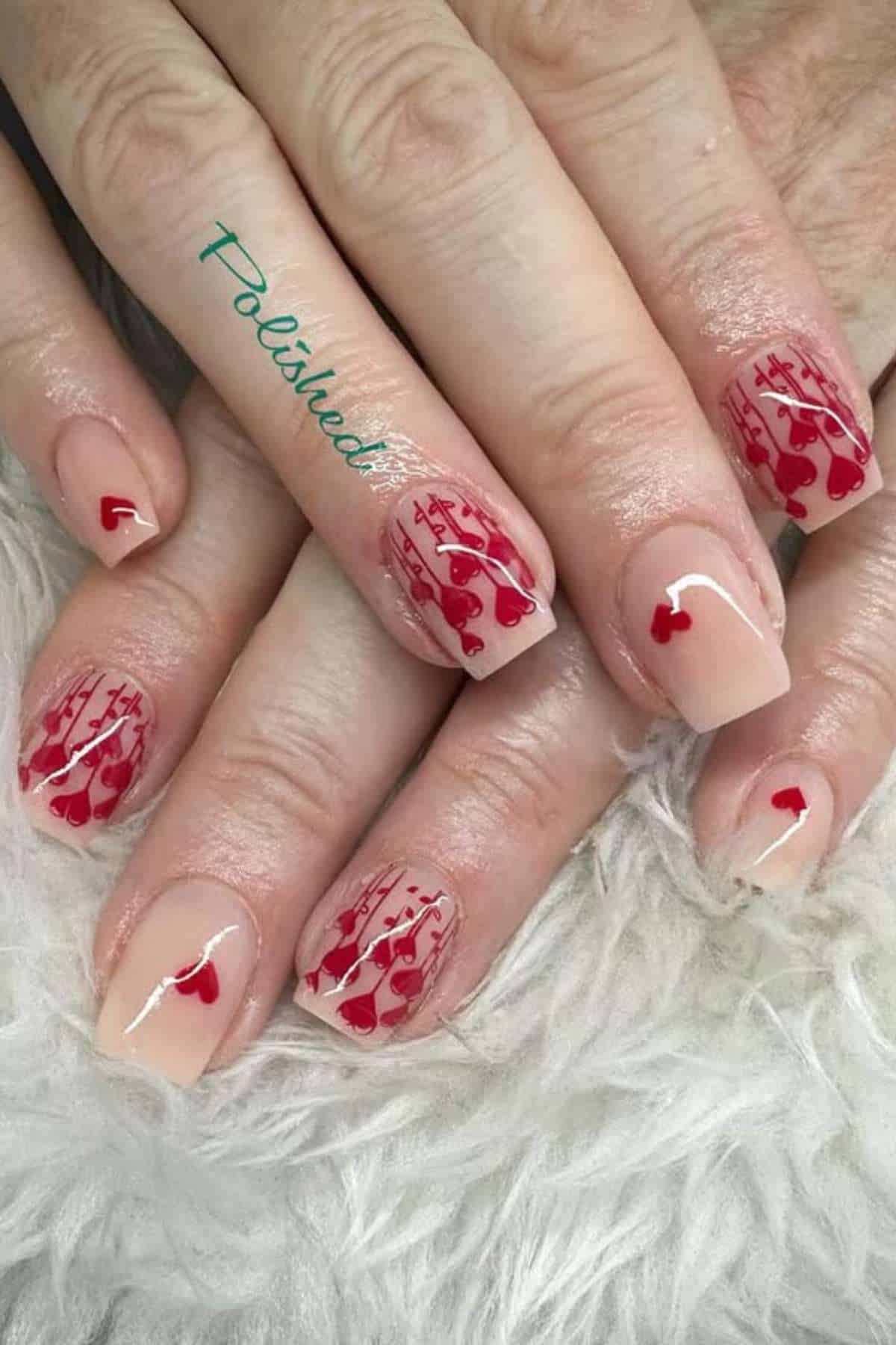 Close-up of hands with nude-colored nails featuring red heart and line nail art, displayed on a soft white surface.