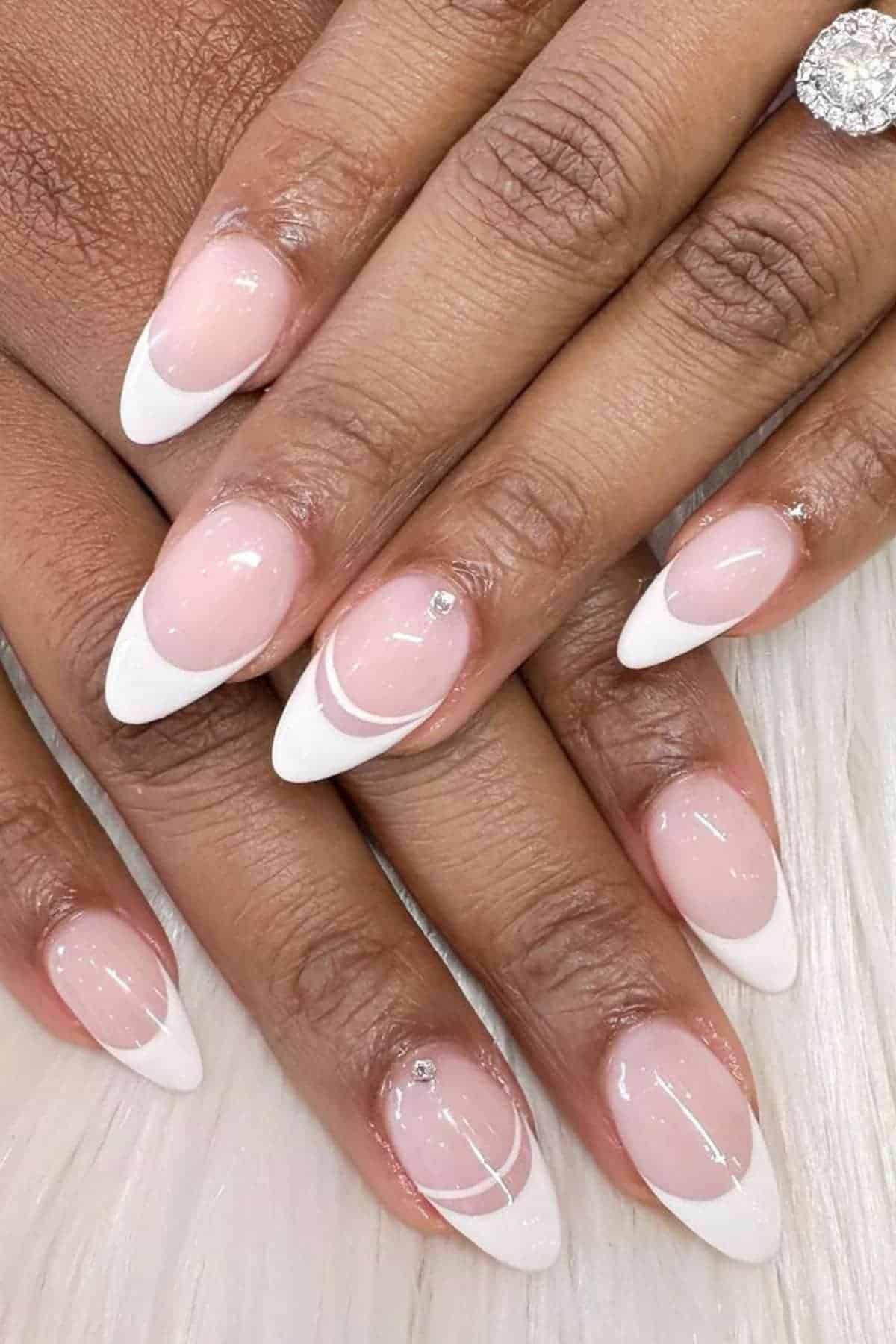 Hands with almond-shaped French tip nails featuring white tips, delicate white line designs, and small rhinestone accents on a light pink base.