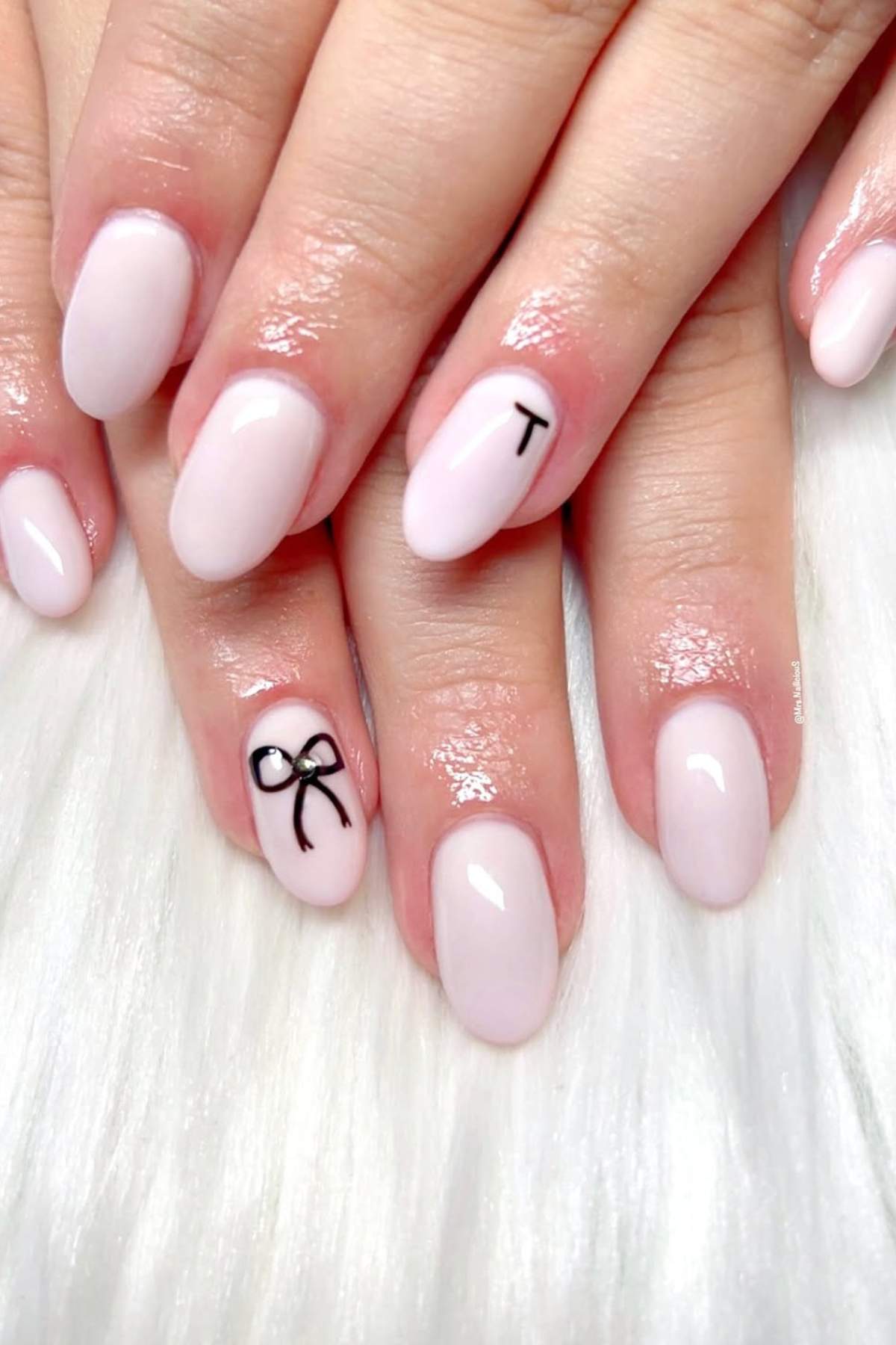 Hands with oval-shaped, pale pink nails; the ring finger has a small black bow design, and the index finger has a black letter "T.
