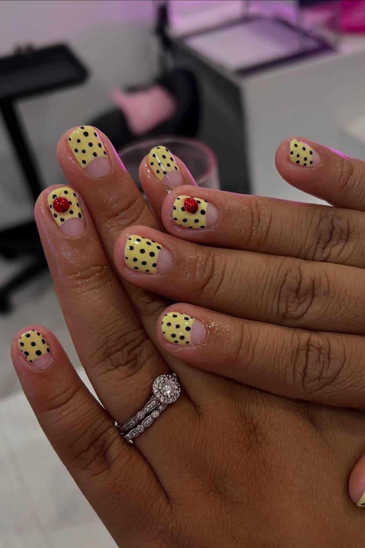 Close-up of hands with yellow nails featuring black polka dots and small red ladybug designs; person is wearing a silver ring with a large stone.