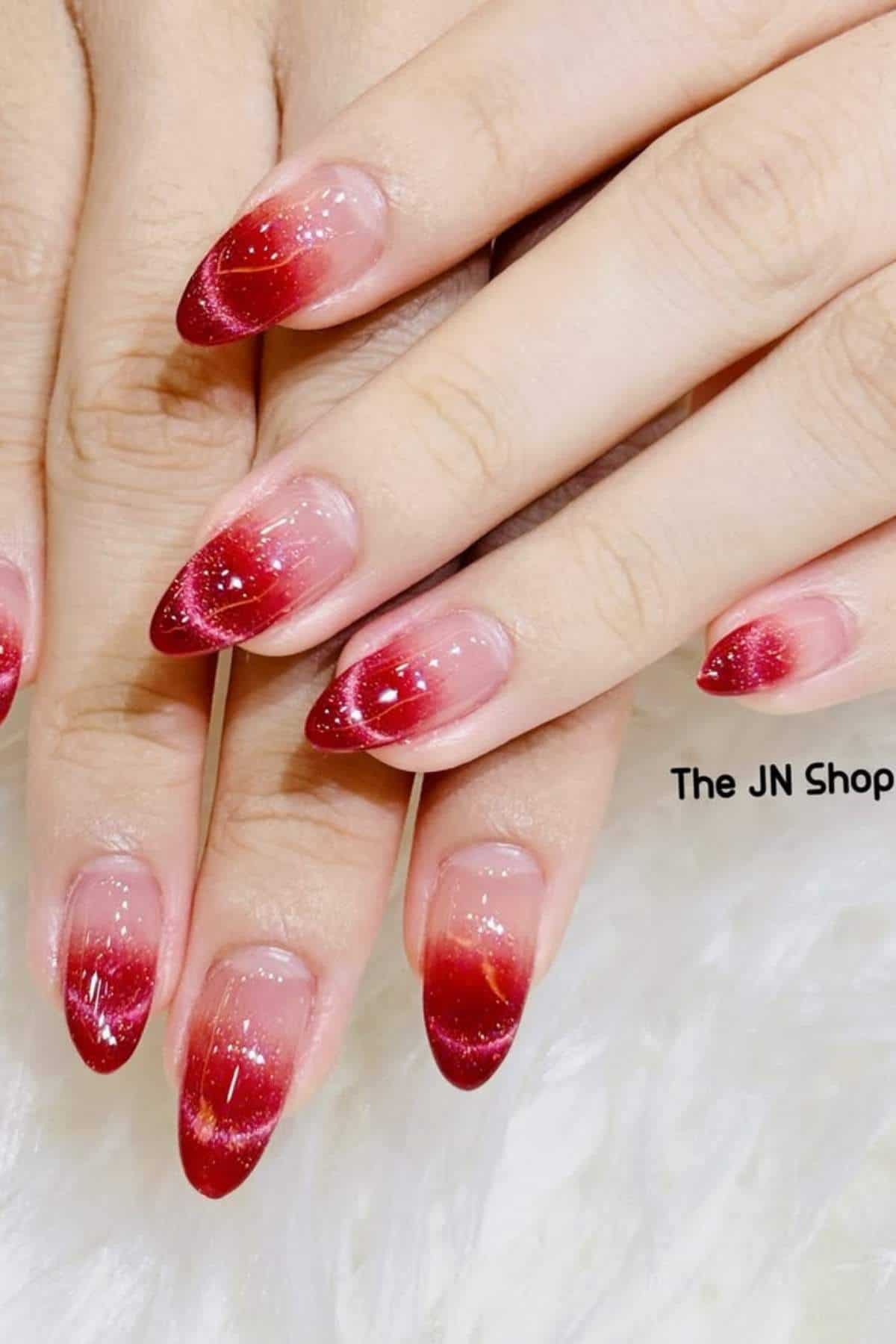 Hands with almond-shaped nails featuring a red glitter ombre design, placed on a white surface. The text "The JN Shop" is partially visible.