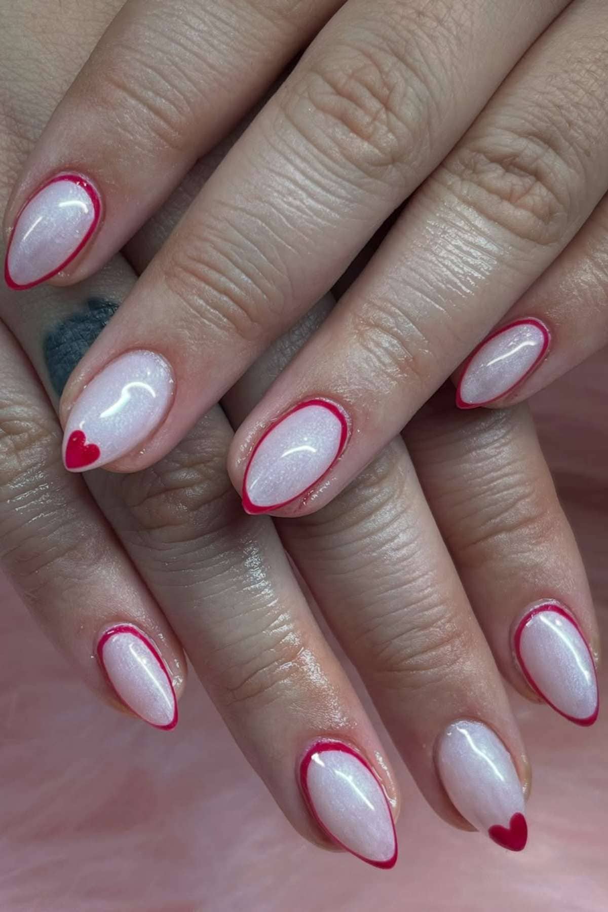 Close-up of hands with almond-shaped nails painted pearly white, featuring thin red outlines and small red heart accents near the tips.