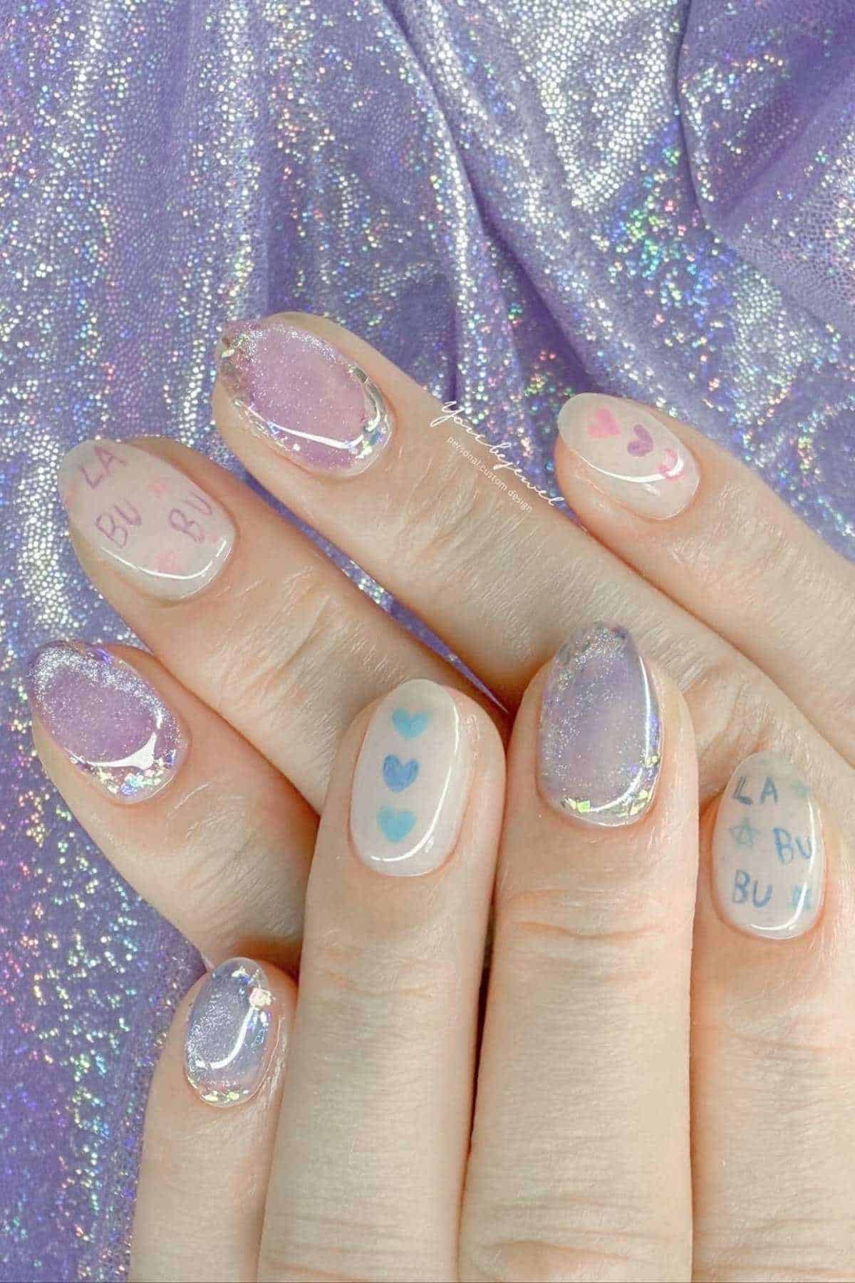 A hand with long, oval nails featuring pastel purple shimmer polish, blue and pink heart designs, and "LA BU BU" text accents on a shimmery lavender fabric background.