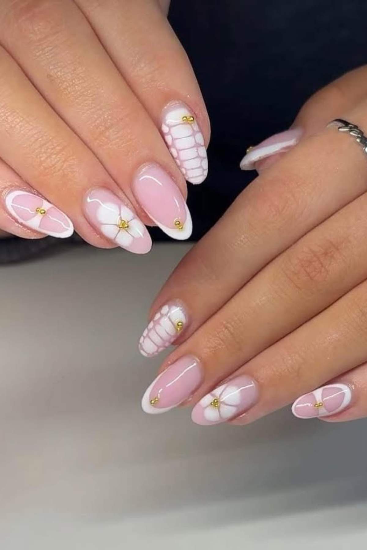 Close-up of hands with almond-shaped nails featuring a pink base, white floral and geometric designs, and small gold bead accents.