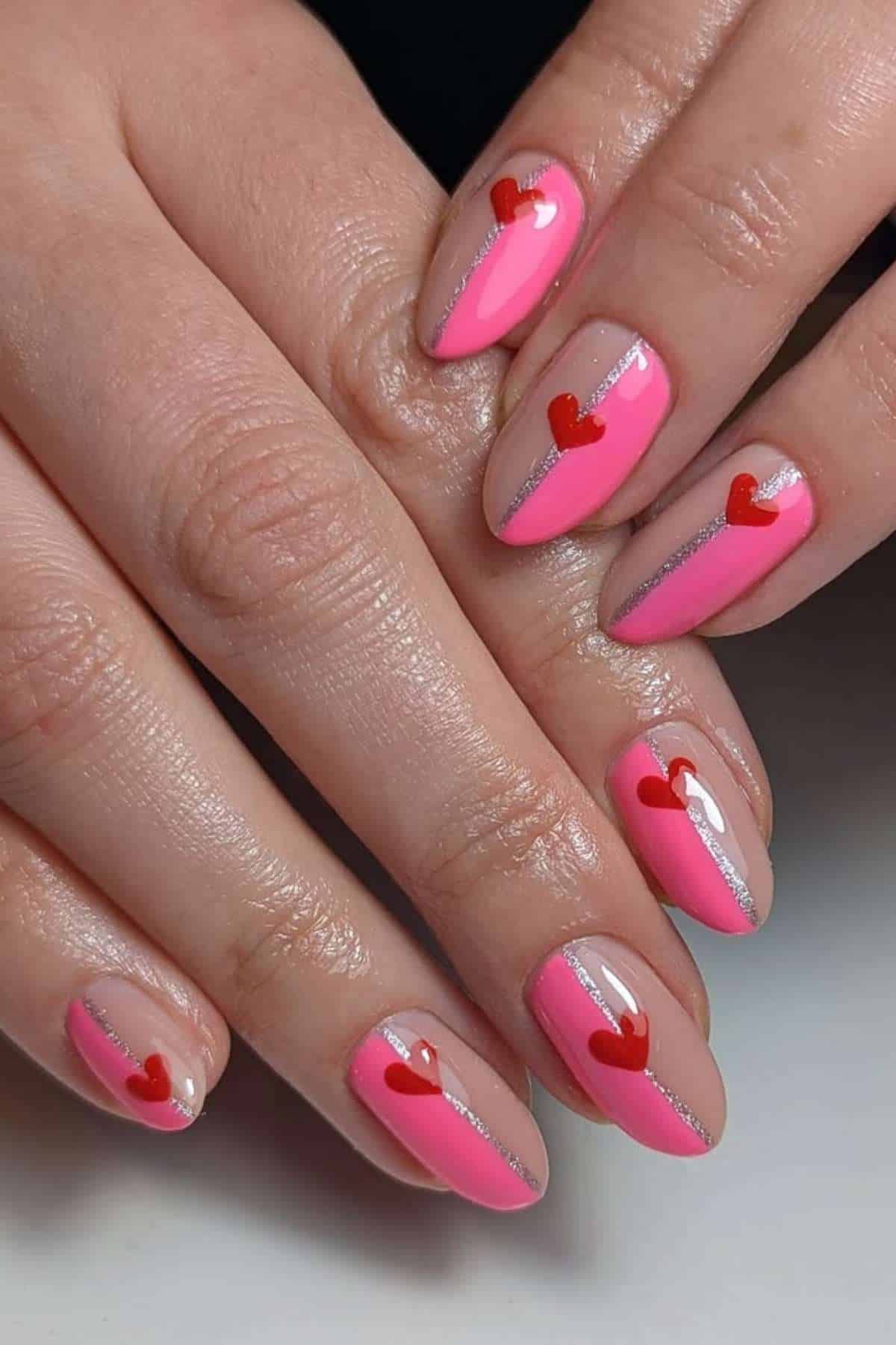 Hands with manicured nails featuring pink polish, a silver diagonal stripe, and a small red heart design on each nail.