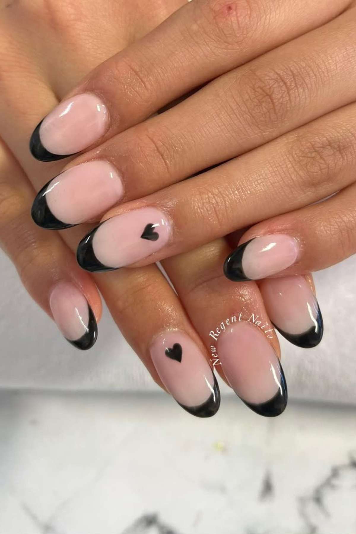 Hands with almond-shaped nails featuring a light pink base, black French tips, and small black heart designs on two nails.