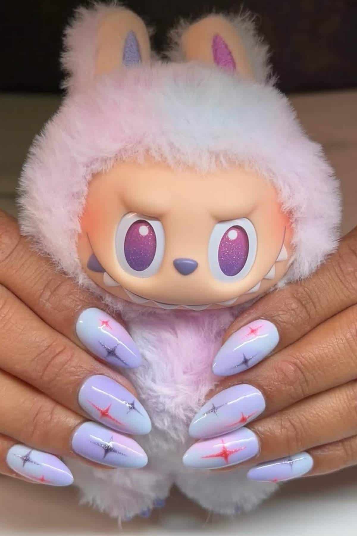 Hands with pastel blue nails featuring pink star designs hold a plush bunny toy with purple eyes and a fluffy pastel-colored hood.