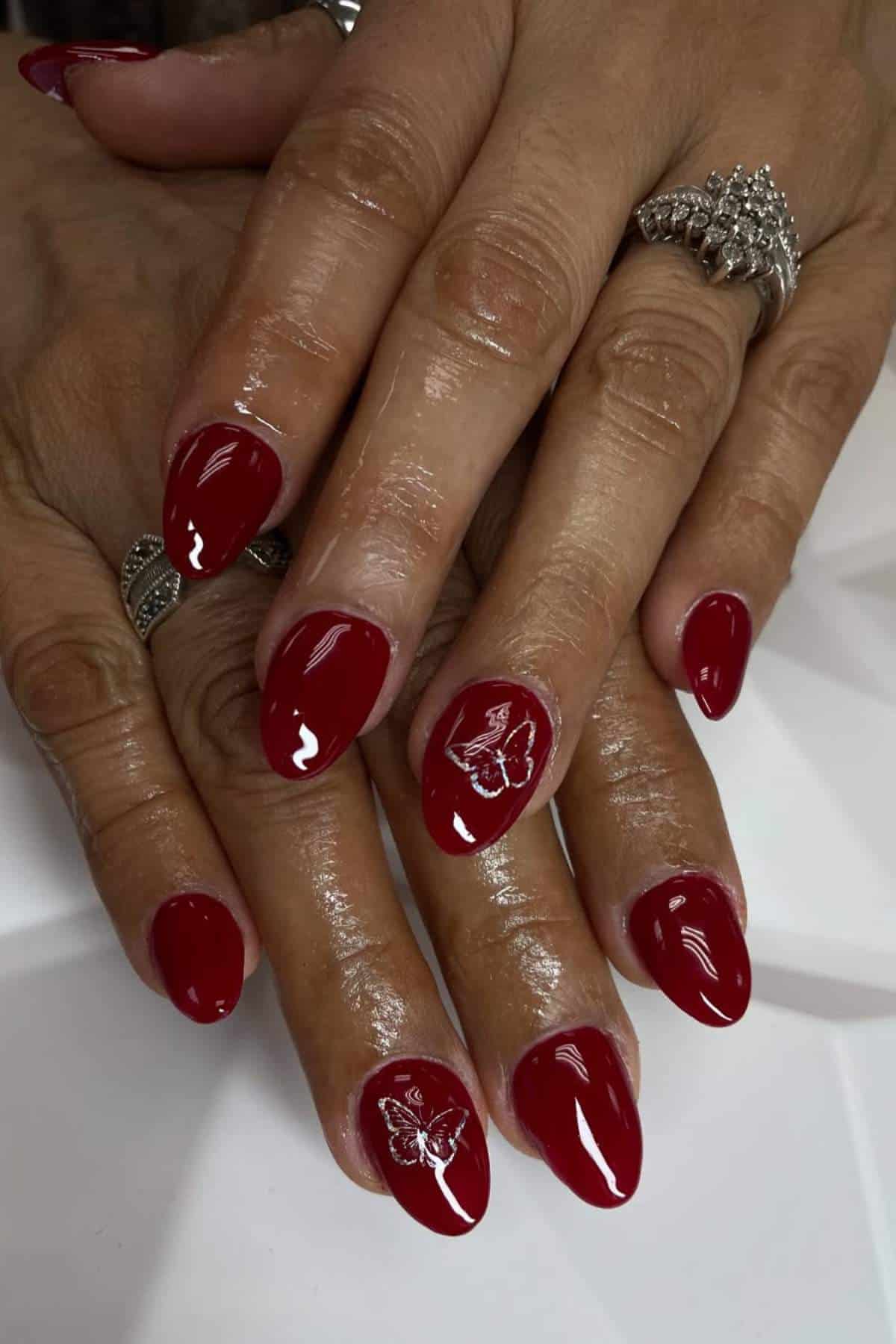 Hands with shiny red almond-shaped nails, featuring a delicate white butterfly design on the ring fingernails. The person is wearing silver rings.
