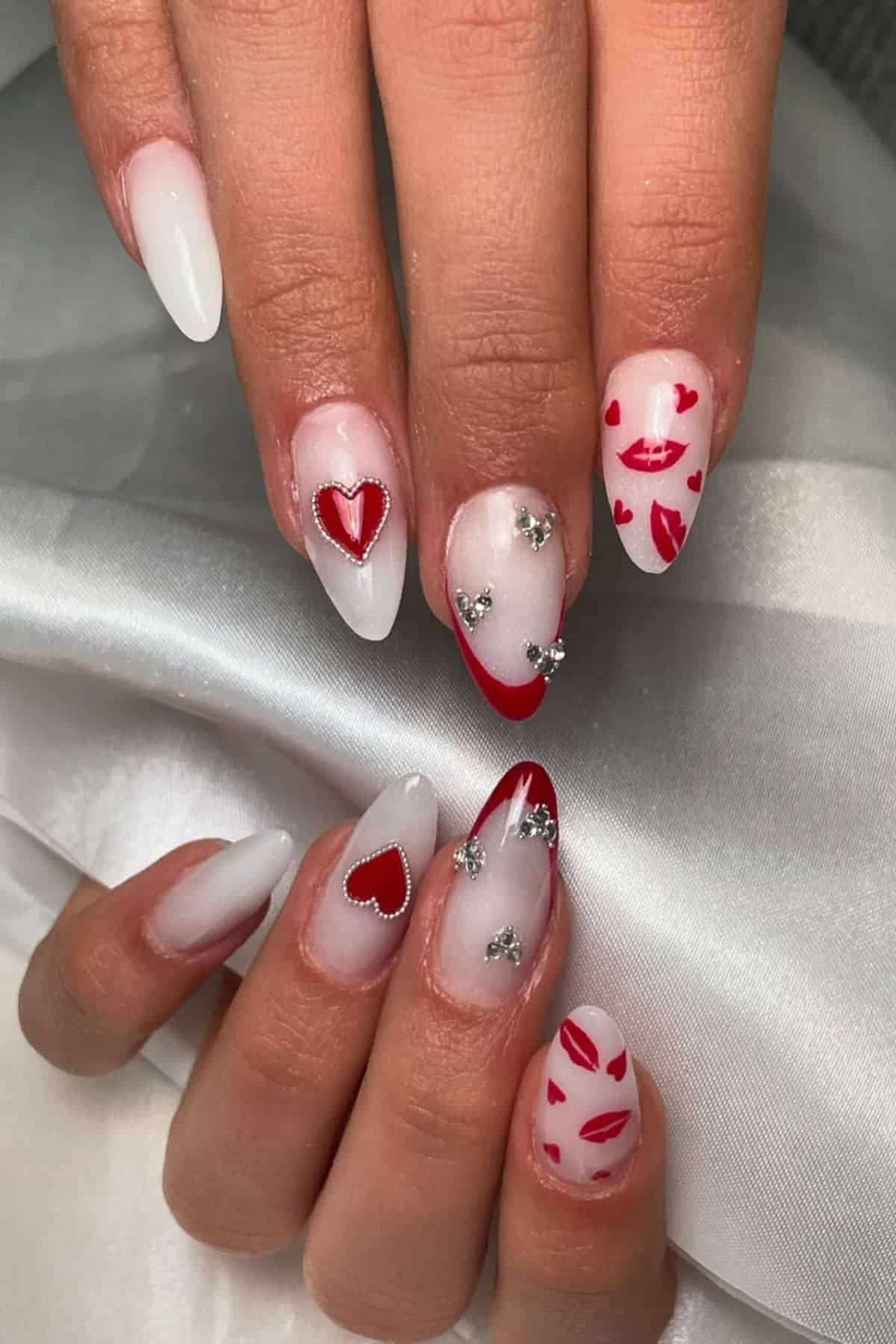 Hands with almond-shaped nails featuring red hearts, lips, silver rhinestones, and red-tipped French manicure designs on a white satin background.