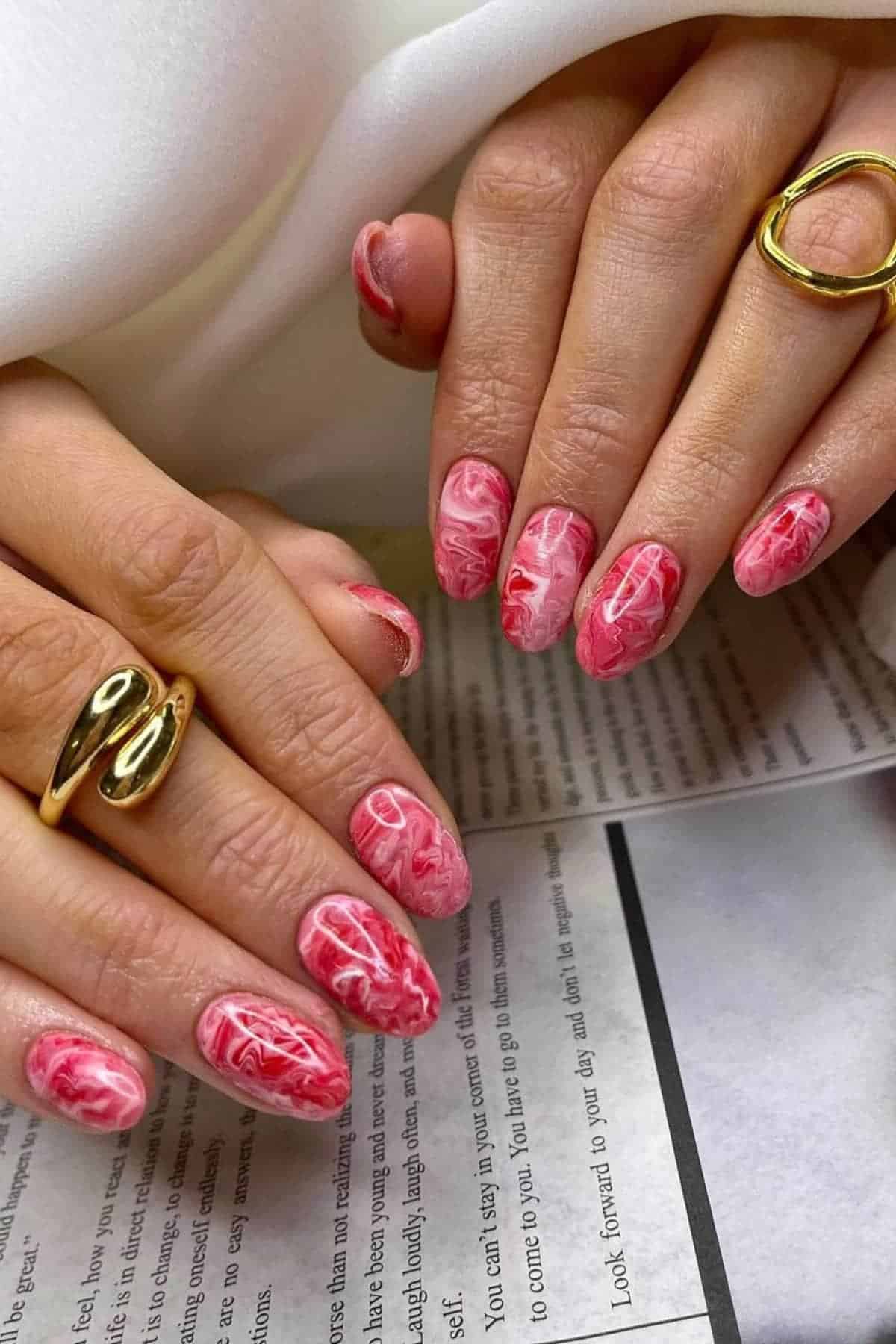 Hands with light red and white marbled nail art, wearing gold rings, resting on a newspaper or printed paper.