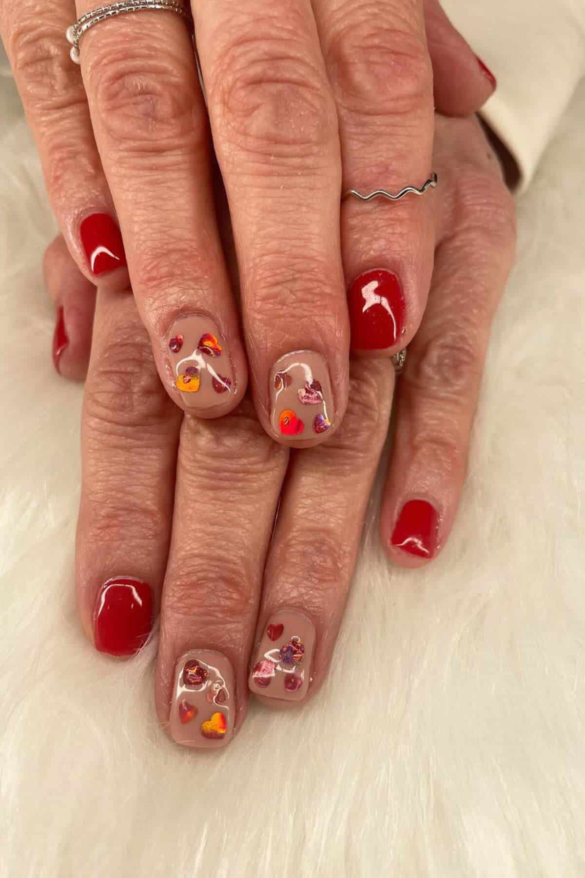 Hands with red nail polish and accent nails featuring colorful heart designs, displayed on a white, fuzzy surface.