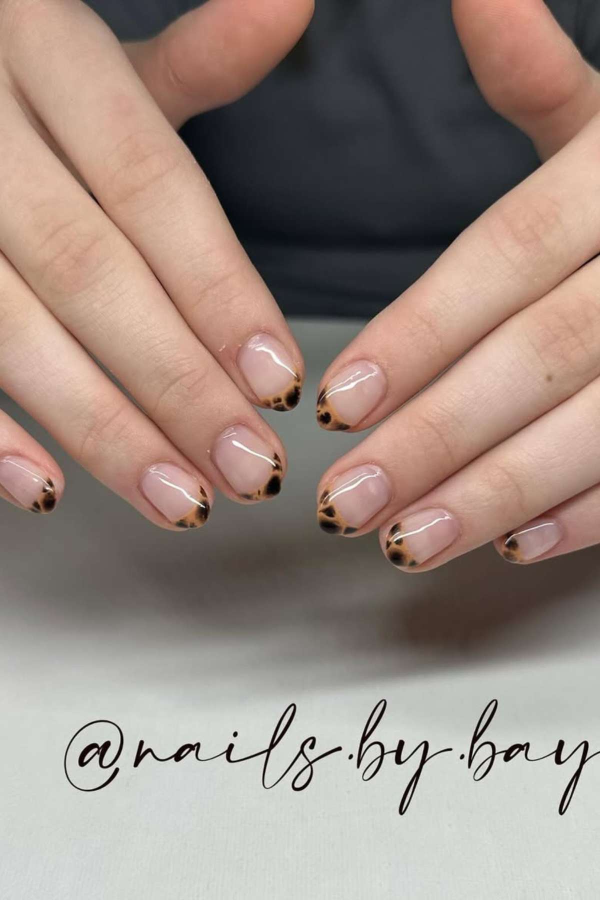 Close-up of hands with short nails featuring a clear base and a brown and black tortoiseshell pattern on the tips. The text "@nails.by.bay" is overlaid at the bottom.