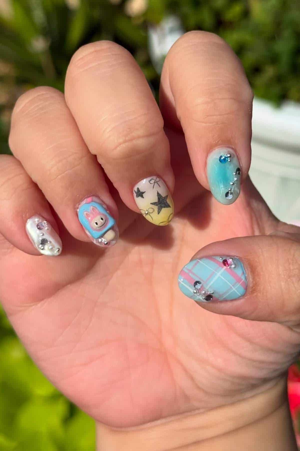 A hand with a mixed nail art design featuring pastel colors, 3D embellishments, a cartoon bunny, stars, ombre blue, and a blue plaid pattern.