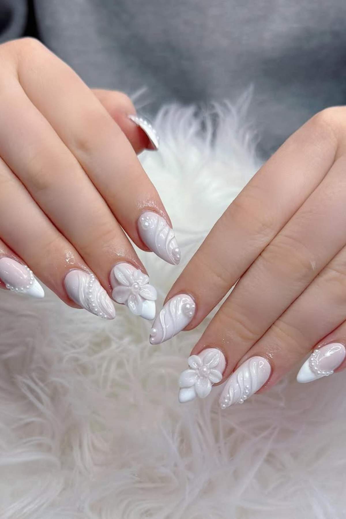 Hands with long, pointed nails featuring white 3D floral and swirl designs, displayed over a fluffy white surface.