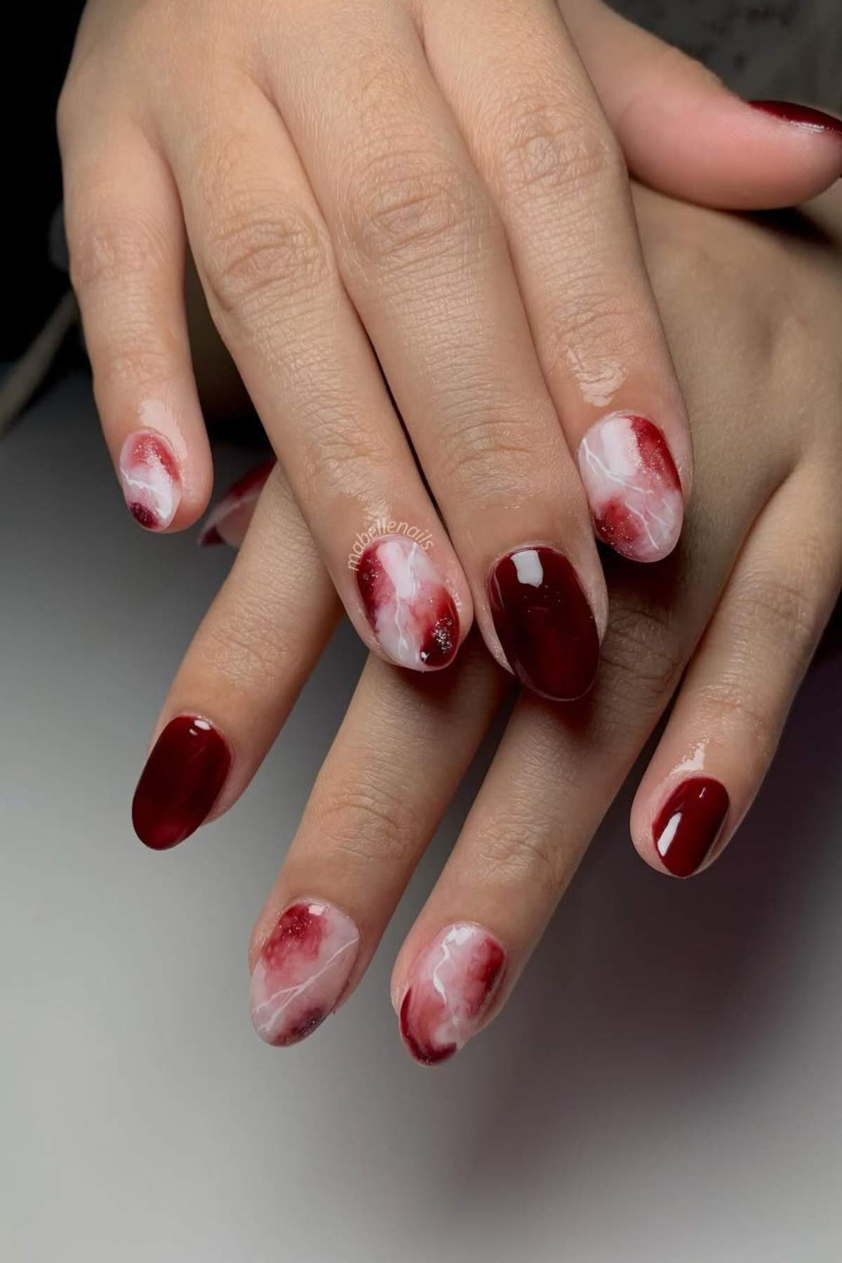 Hands with manicured nails featuring a mix of deep red polish and marble-like red and white designs on a few accent nails.
