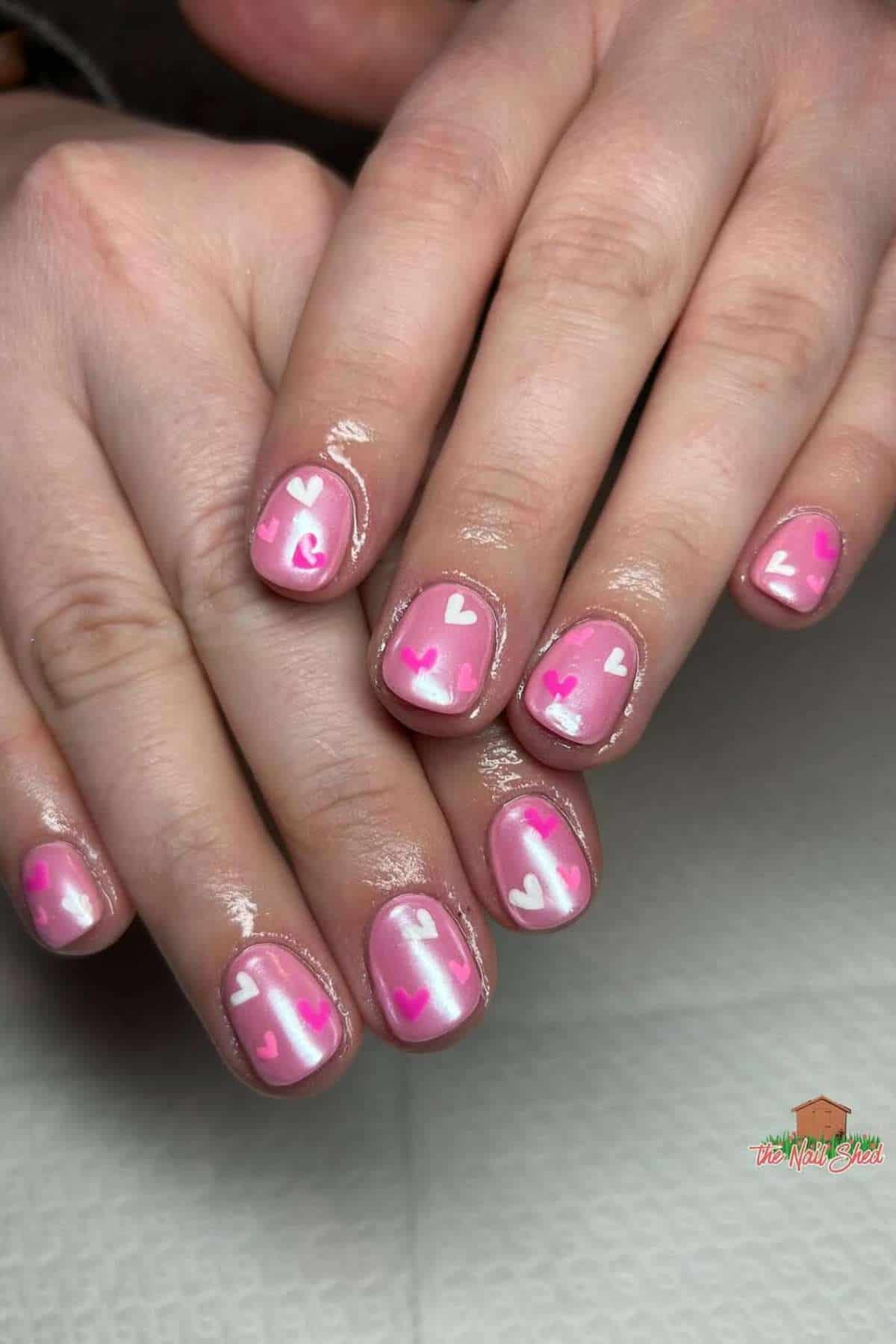 Hands with short, pink-manicured nails featuring white and pink heart designs on each nail. The nails have a glossy finish.