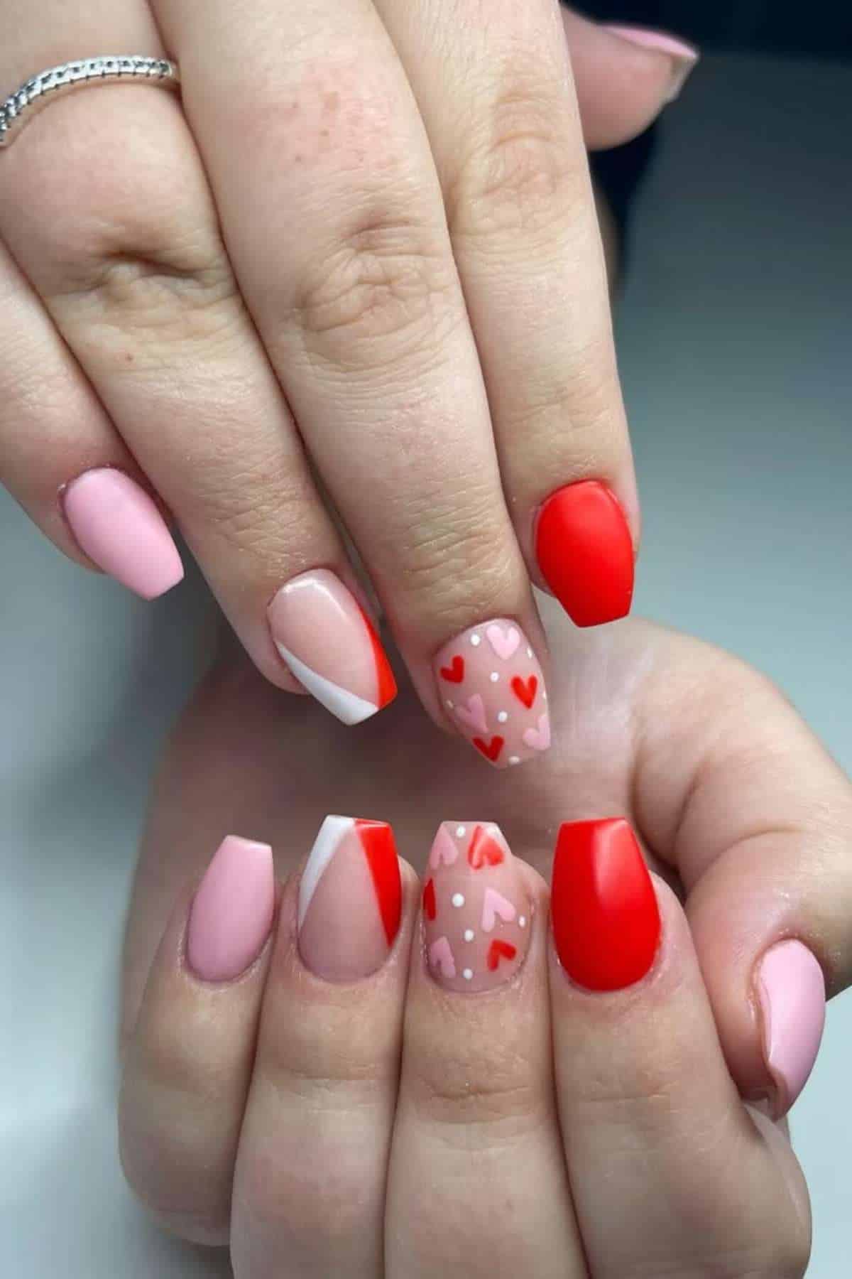 A close-up of hands with manicured nails featuring pink, red, and white designs, including small red hearts and geometric patterns.