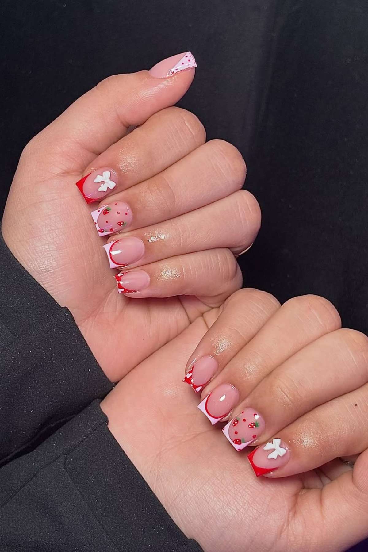 Two hands with manicured nails featuring red French tips, small flower designs, polka dots, and a nude pink base, against a black background.