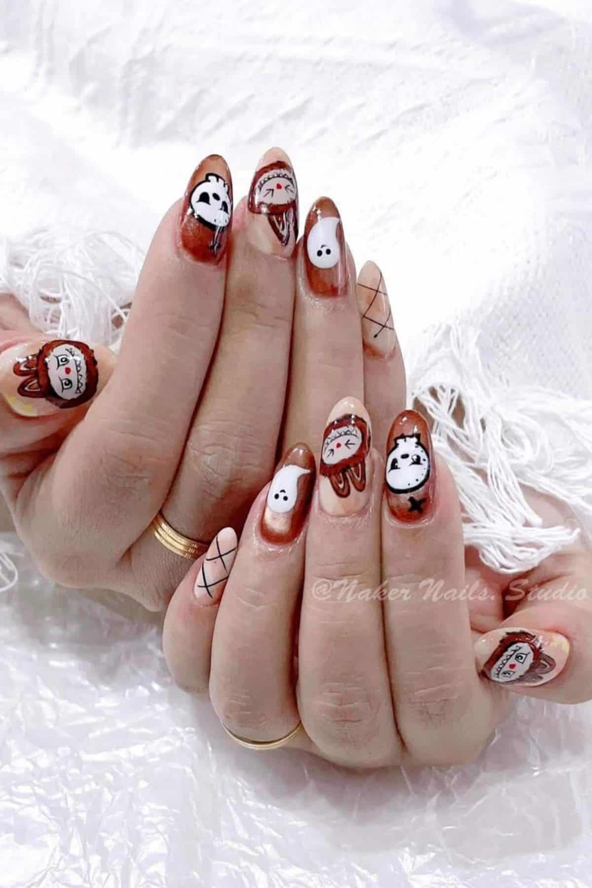 Hands with long nails painted in brown, white, and red, featuring Halloween-themed designs such as ghosts, pumpkins, and stitched faces, resting on white fabric.