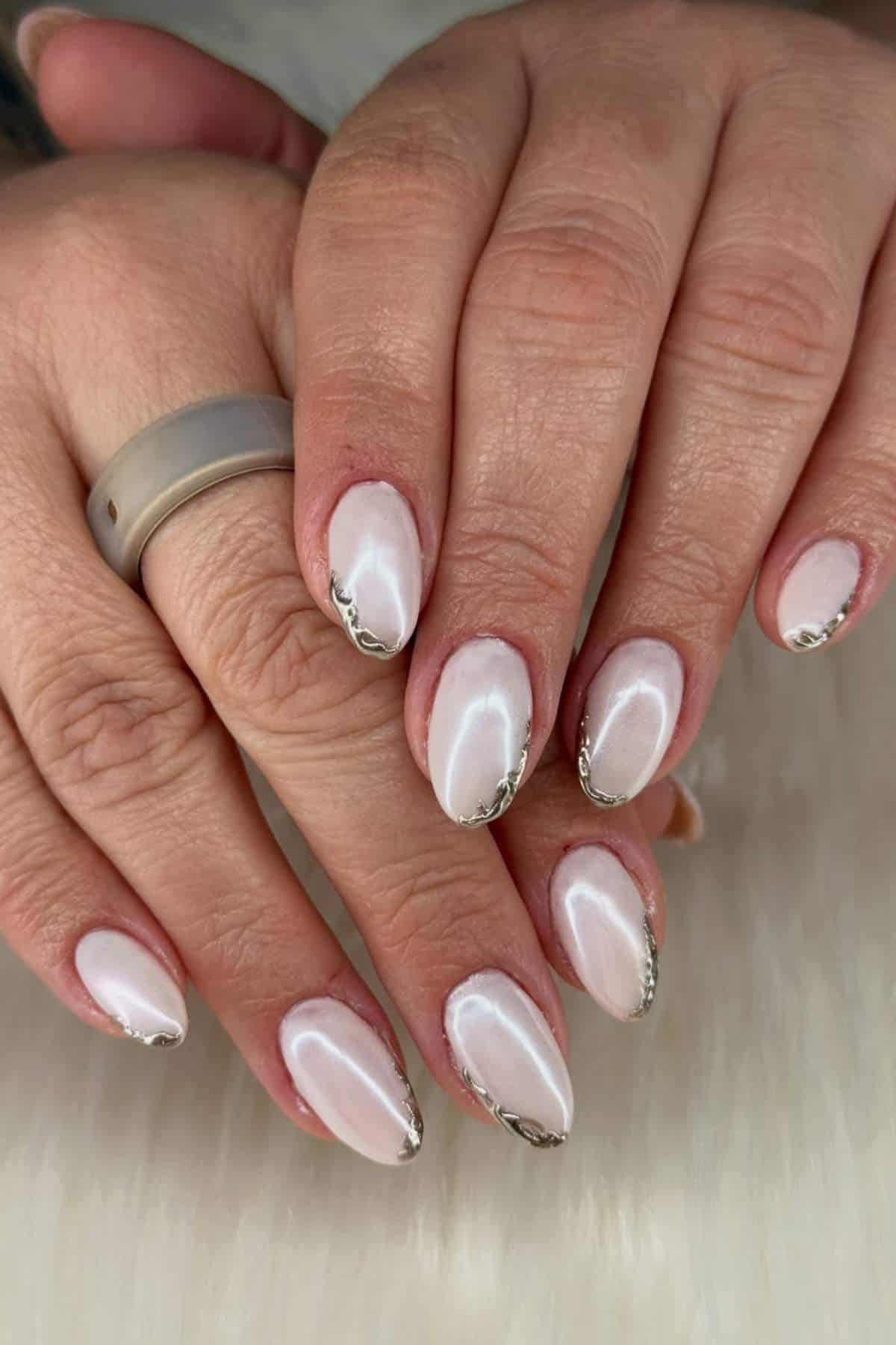 Close-up of hands with almond-shaped nails, painted in a pearly white polish and accented with metallic silver tips. A matte silver ring is worn on one finger.