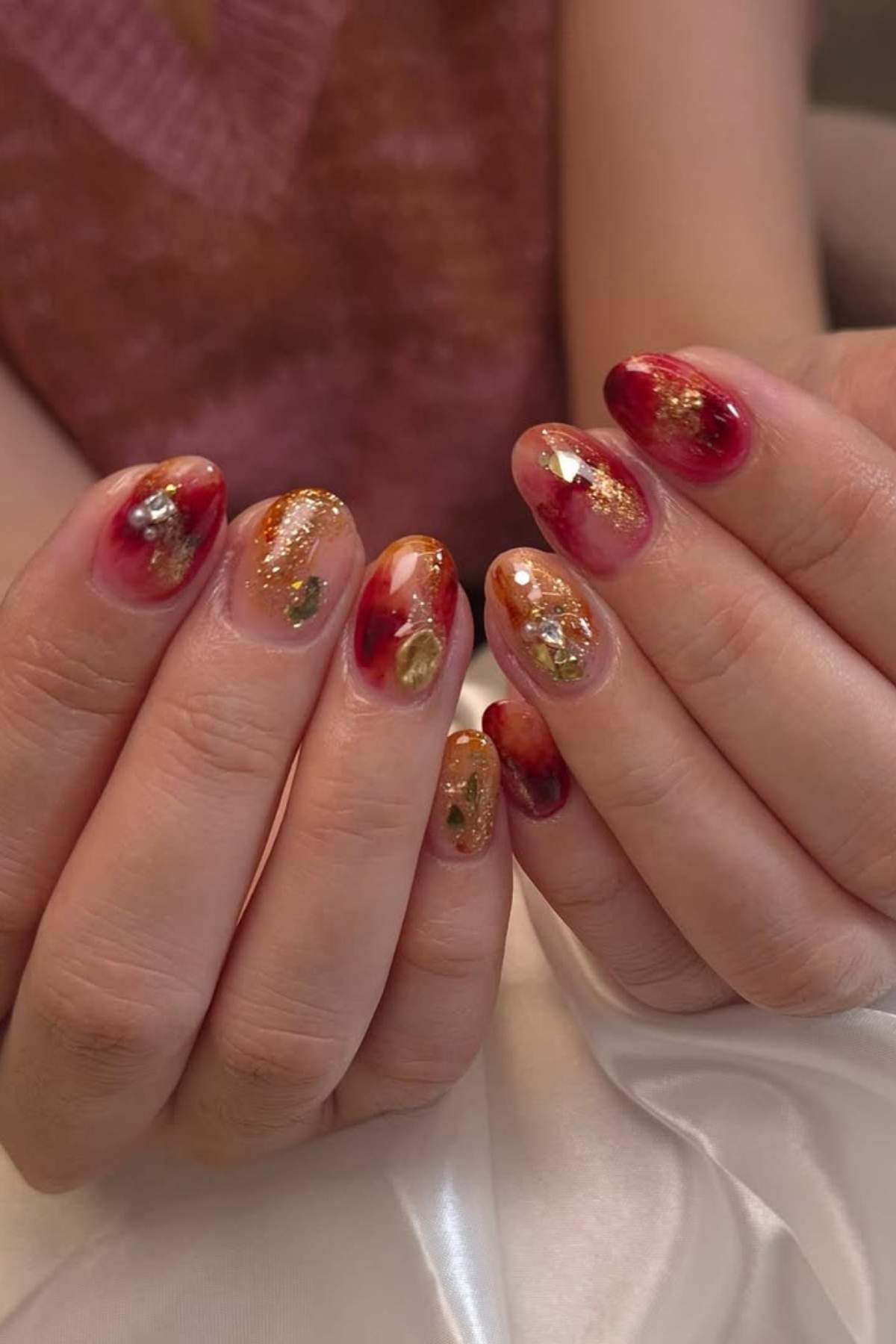 Hands with almond-shaped nails featuring a glossy red and gold marbled design with glitter and foil accents.