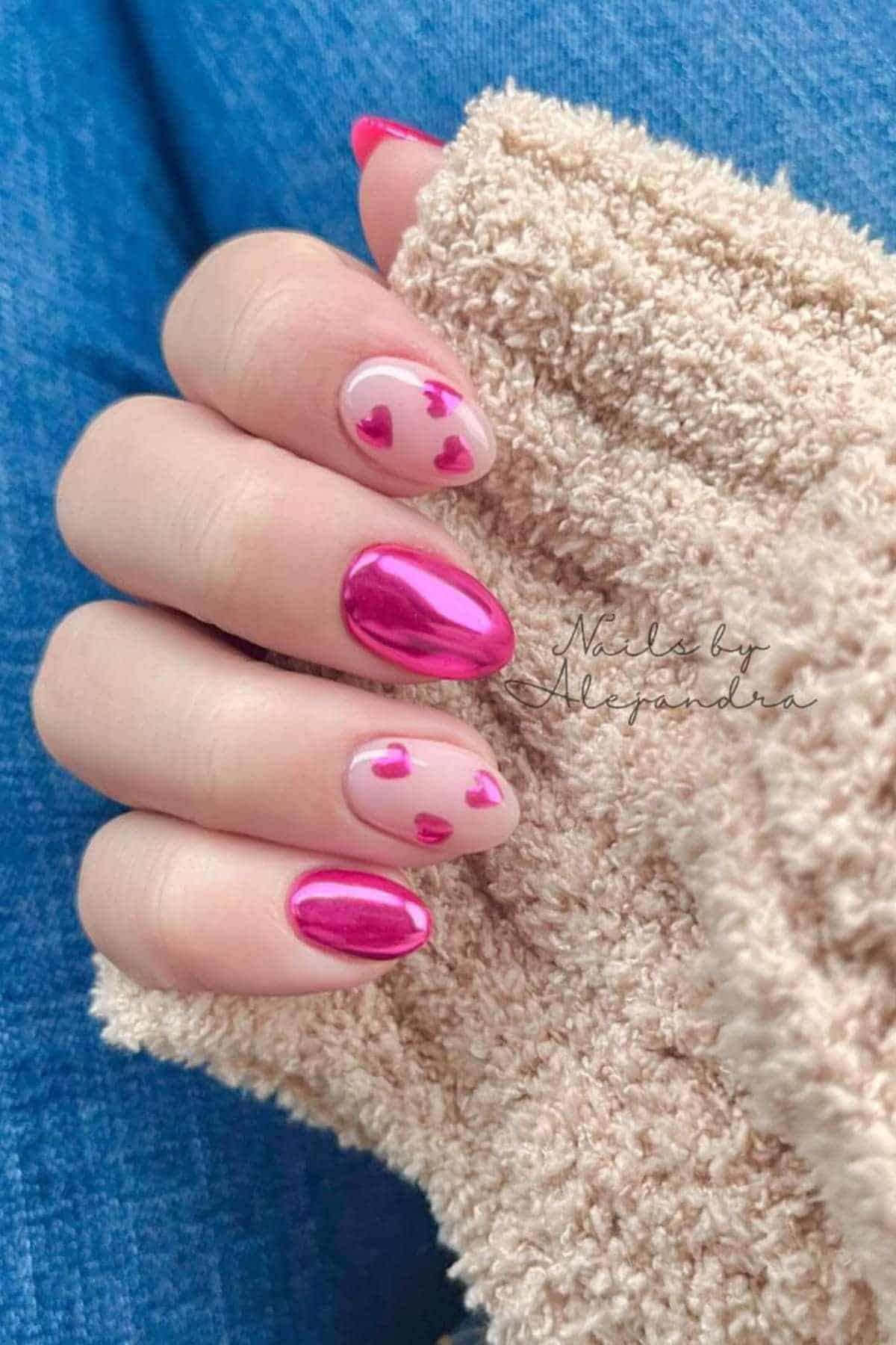 A hand with pink metallic and nude nails, some featuring small pink heart designs, is shown against a beige textured fabric and blue jeans background.
