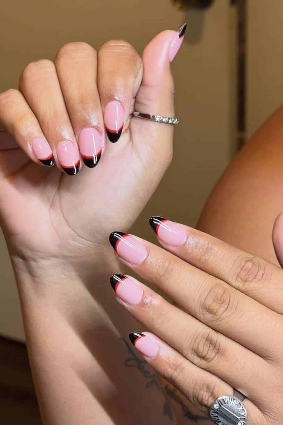 Hands with almond-shaped nails featuring a pink base and black French tips, accented with a thin red line. One hand wears a silver ring.