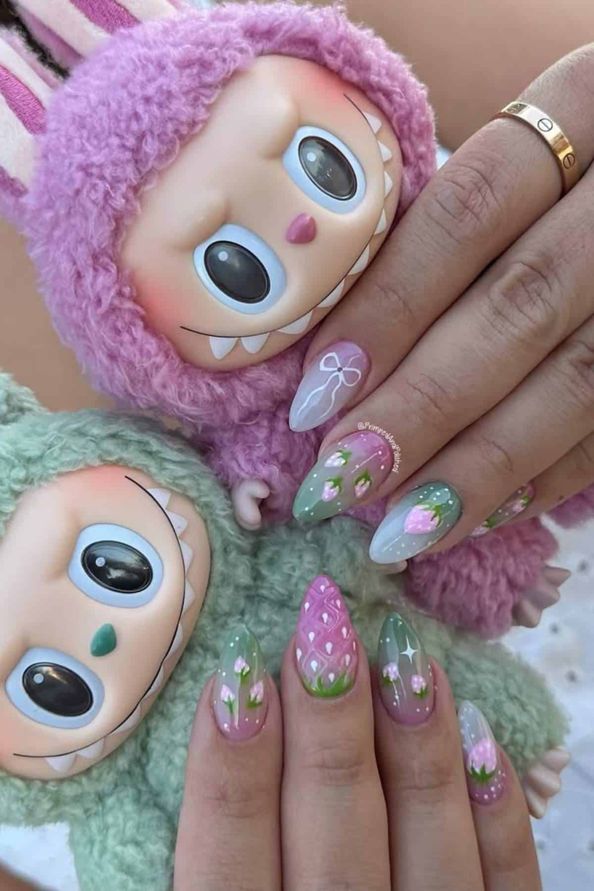 Hands with pastel green and pink strawberry-themed nail art hold two plush dolls with large eyes, one in a pink suit and the other in a green suit.