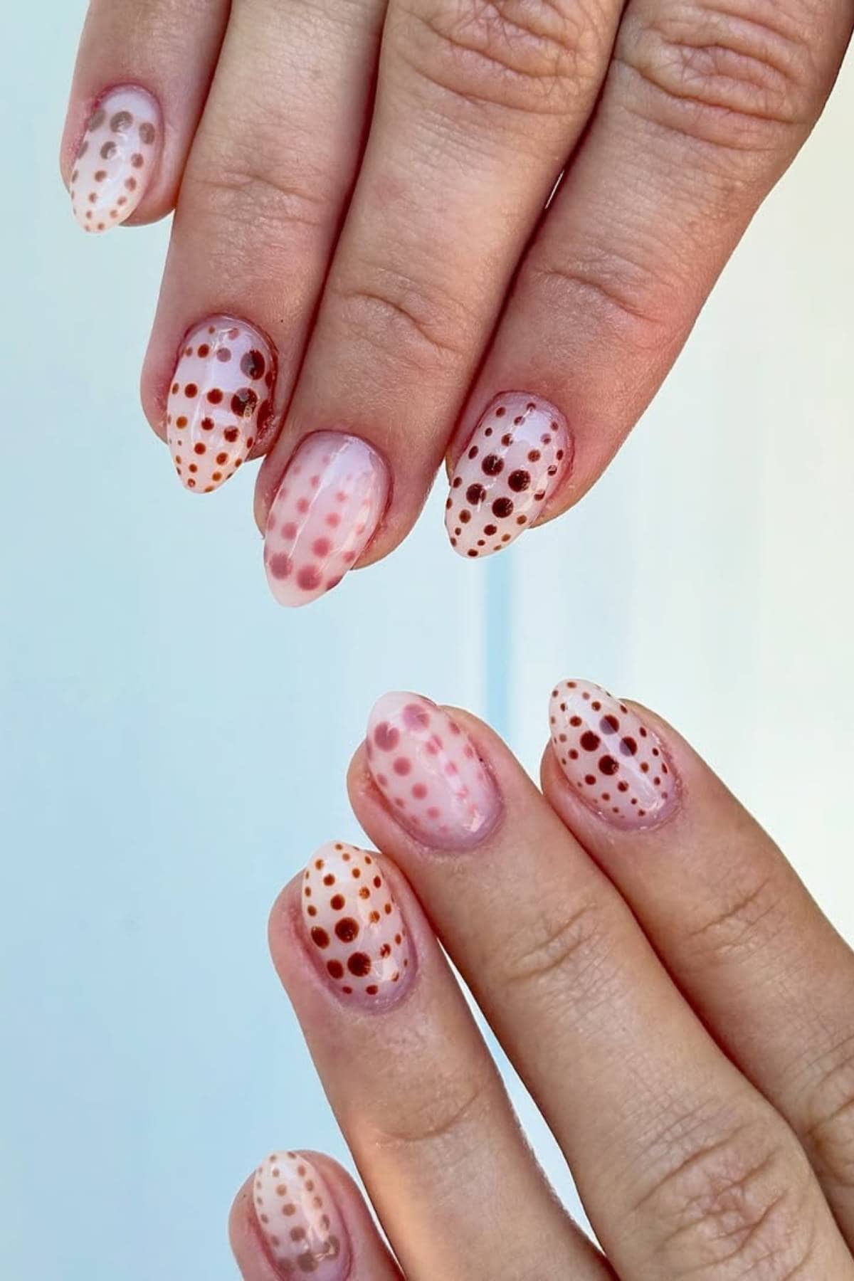 Close-up of hands with short, oval nails painted in a translucent nude color and decorated with rows of red, brown, and orange polka dots.